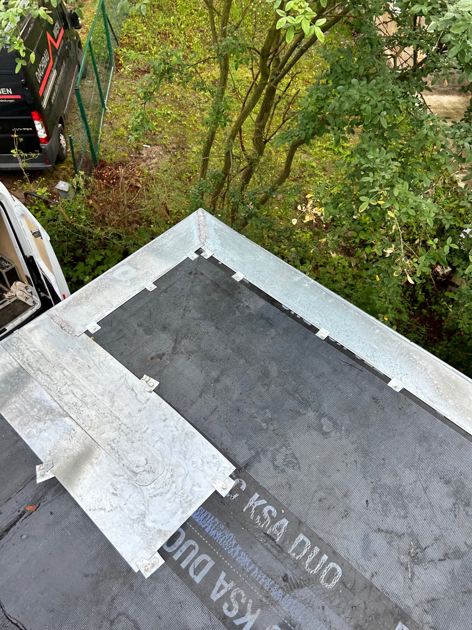 Widok z góry na fragment dachu z nową, czarną membraną hydroizolacyjną KSA DUO, obróbki blacharskie z jasnego metalu i częściowo zamontowaną blachą trapezową, w tle zieleń drzew i samochód firmy...