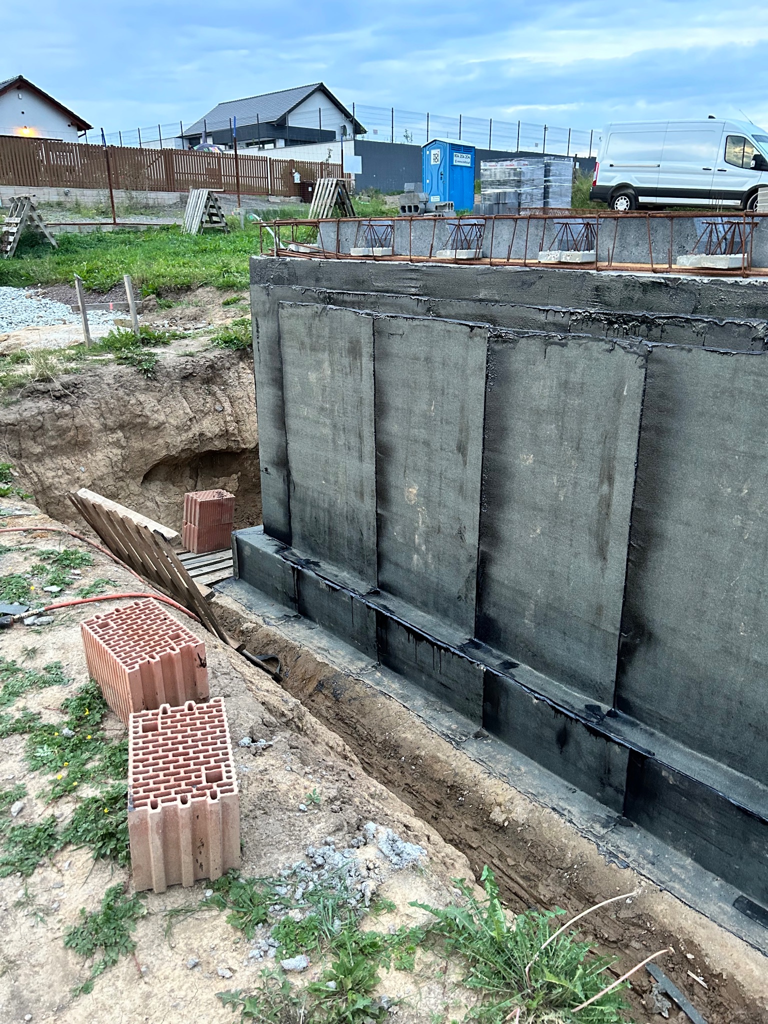 Izolacja pionowa fundamentów budynku w trakcie budowy, zabezpieczona czarną folią hydroizolacyjną, widoczne pustaki ceramiczne obok wykopu, w tle dom jednorodzinny i błękitny kontener.