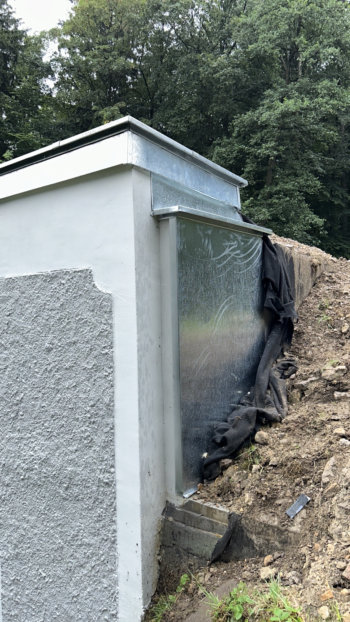 Szara, betonowa konstrukcja z metalowym obiciem i rynną, częściowo zakryta czarną tkaniną, otoczona ziemią i zielenią.