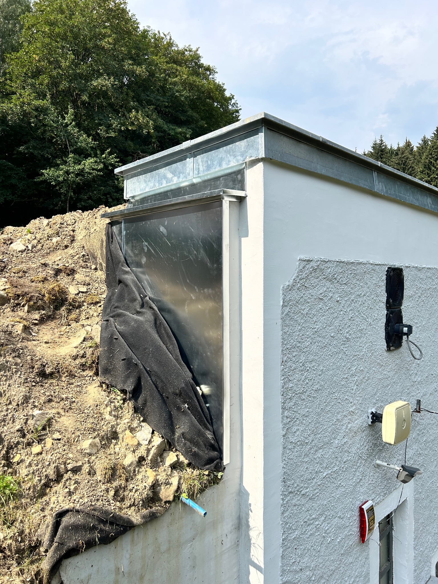 Biały budynek z obróbką blacharską dachu, ziemią i czarną tkaniną przy ścianie, widoczne elementy instalacji na elewacji, w tle drzewa.