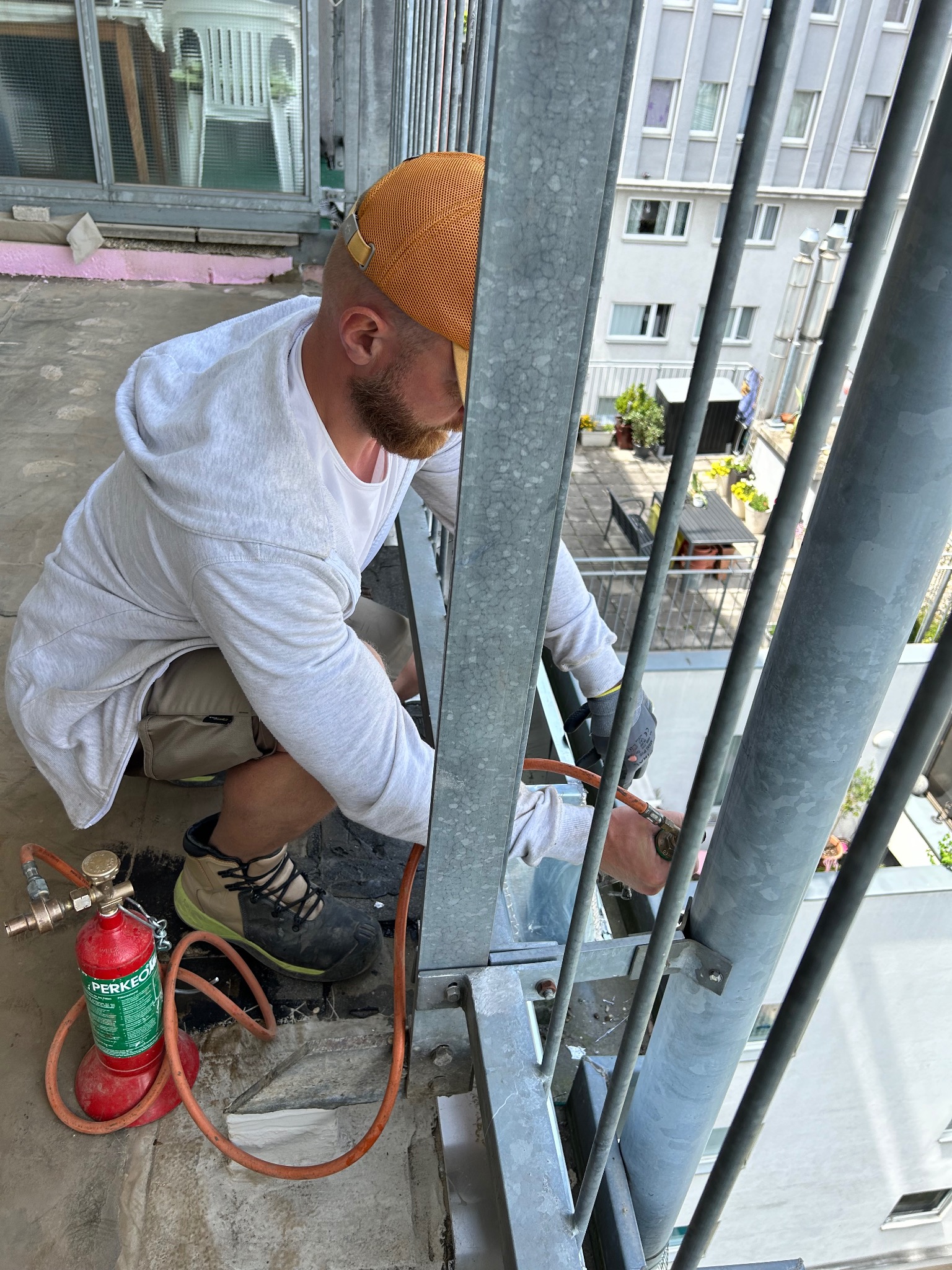 Dekarz w trakcie pracy na balkonie, używający palnika do obróbki blacharskiej, widoczne narzędzia i elementy konstrukcji balkonu.