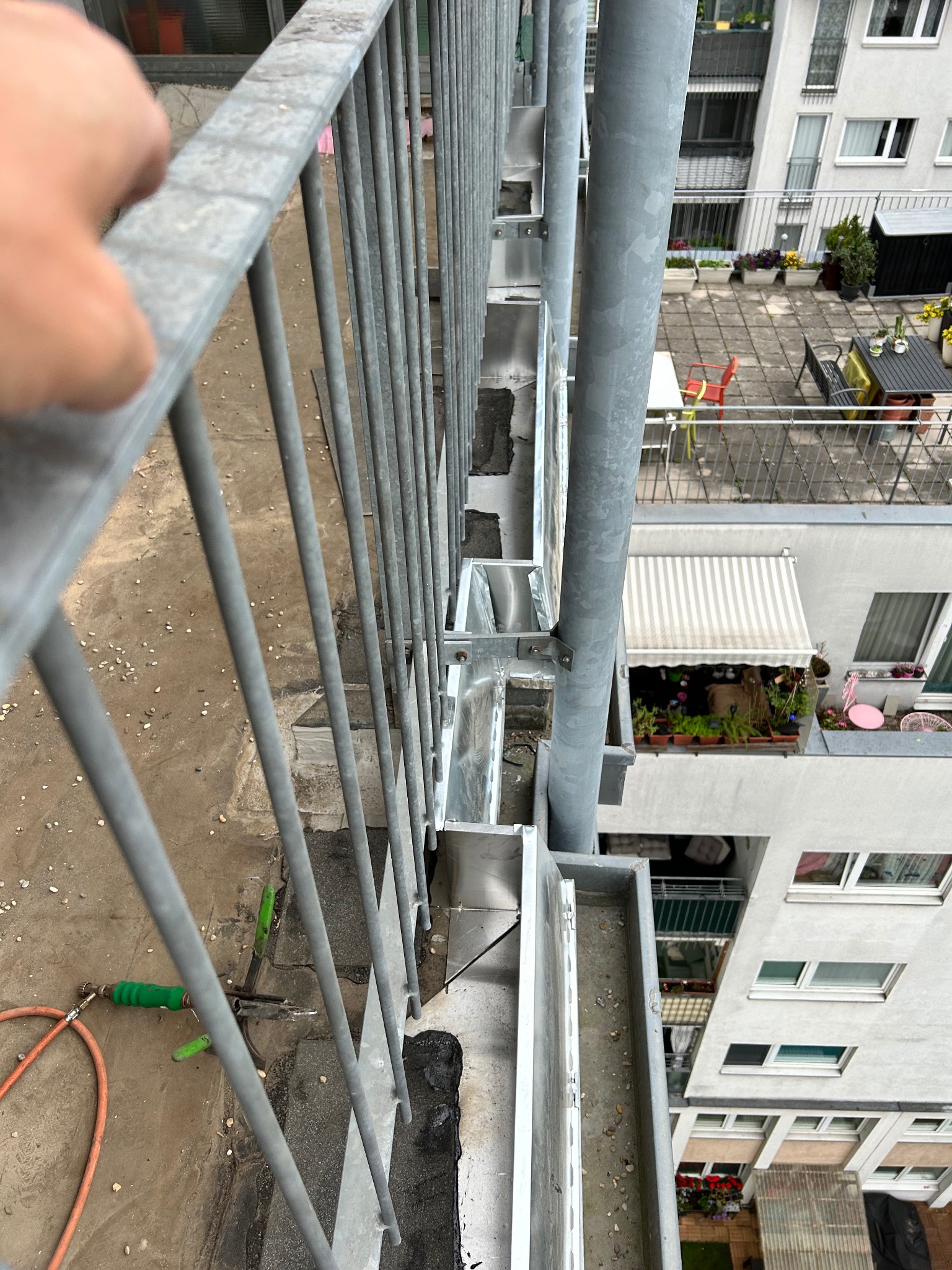 Widok z góry na fragment dachu z zamontowanymi metalowymi rynnami, widoczna ręka na balustradzie, narzędzia dekarskie i fragment elewacji budynku z balkonami.