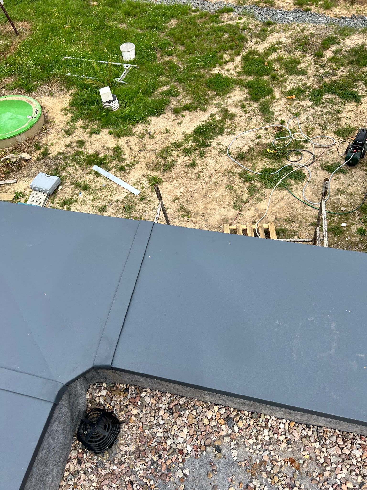 Nowoczesne, grafitowe pokrycie dachu z obróbką blacharską, widok z góry na wykończony narożnik z odpływem wody i żwirem, w tle trawnik i narzędzia.
