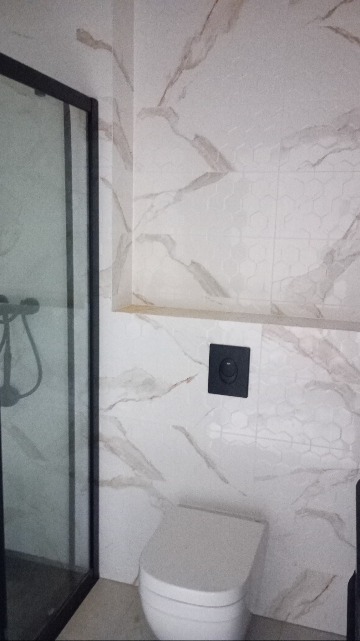 Wykończona łazienka: kabina prysznicowa z czarną ramą, wisząca toaleta i ściana z białych heksagonalnych płytek imitujących marmur z półką i czarnym przyciskiem spłukującym.