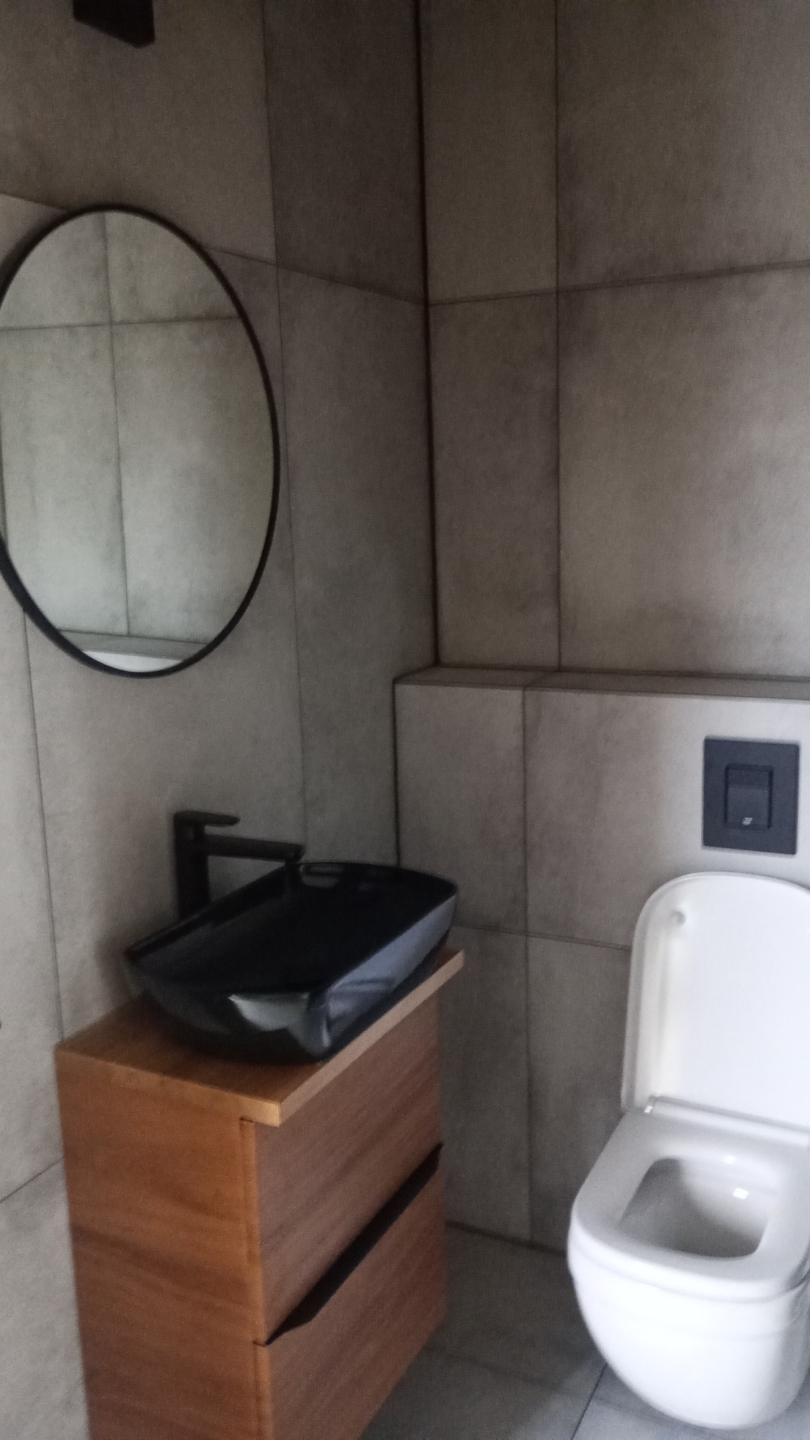 Nowoczesna łazienka z wiszącą, czarną umywalką na drewnianej szafce z szufladami, okrągłe lustro i biała toaleta w tle, szare płytki na ścianach.