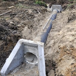 Mini Kop Eryk Kujawa - Rów z betonowymi studzienkami i perforowaną rurą odprowadzającą wodę, otoczony ziemią i drzewami.