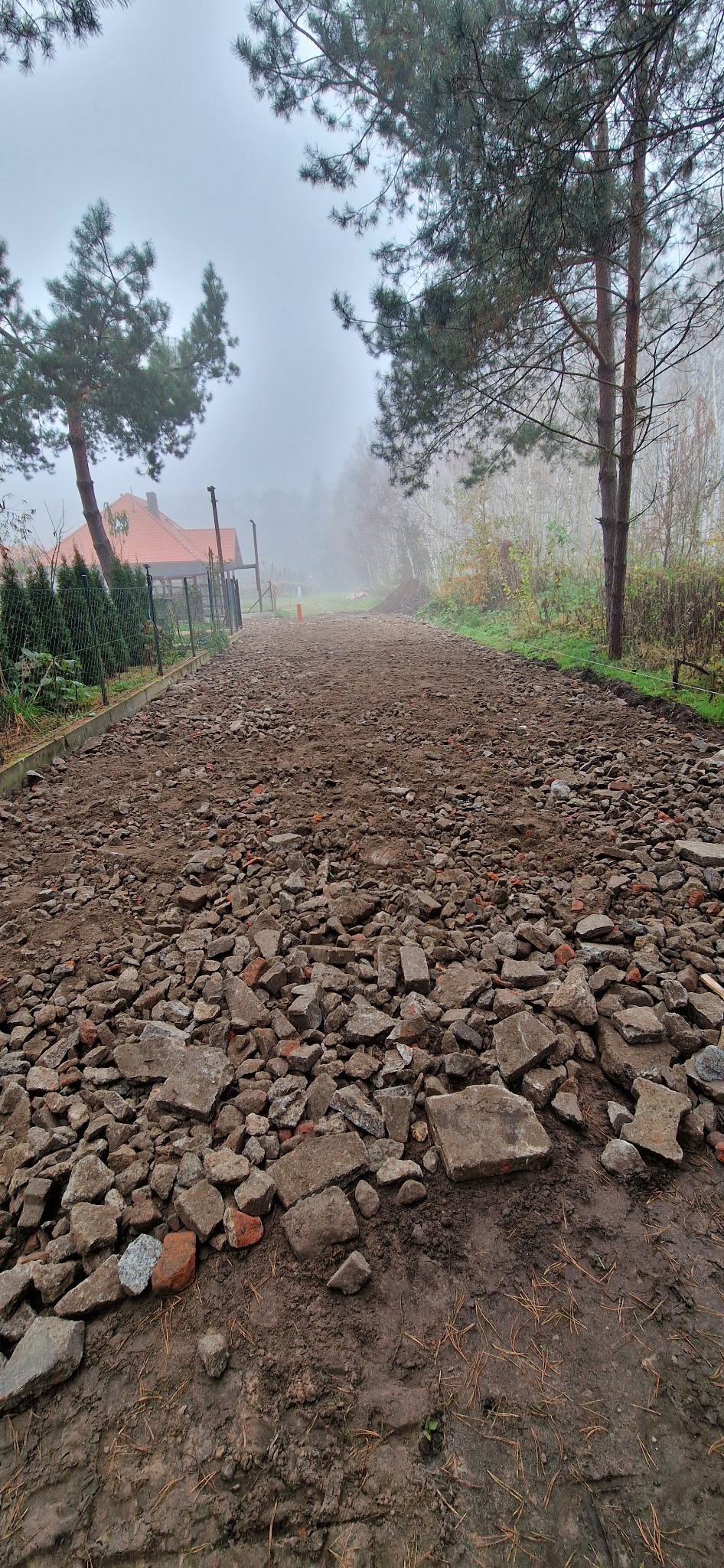 Przygotowanie podłoża pod drogę: gruz i ziemia na działce w mglisty dzień, widoczne drzewa i ogrodzenie w tle.