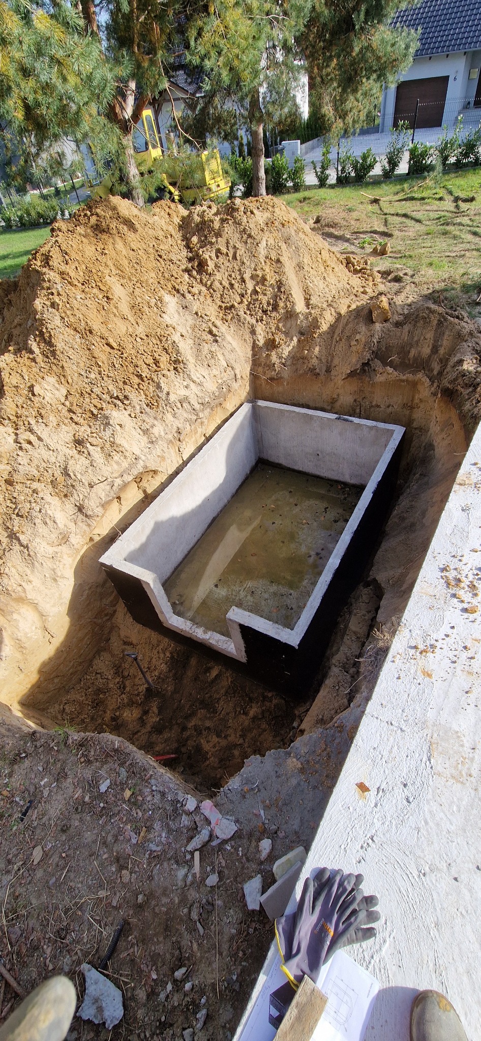 Wykopany dół z betonową konstrukcją w środku, wypełnioną wodą, na tle domu jednorodzinnego i drzew, widoczne rękawice robocze i plany na betonie obok wykopu.