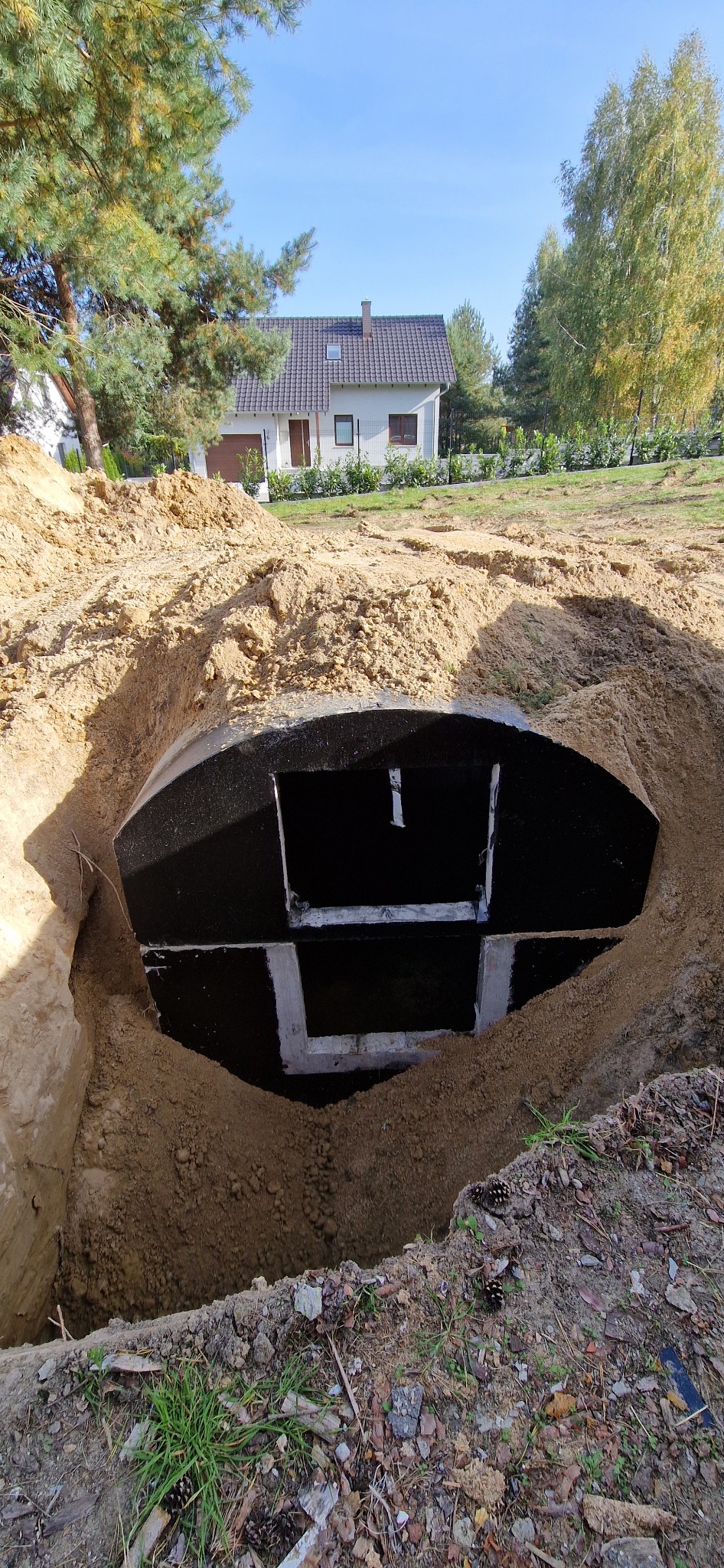 Czarny, betonowy zbiornik szamba częściowo zagrzebany w ziemi, widoczne otwory w górnej części, w tle dom jednorodzinny i drzewa.