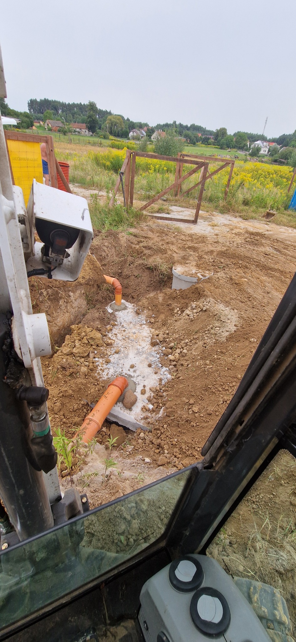 Pomarańczowe rury kanalizacyjne wystające z wykopu, widok z kabiny koparki, w tle krajobraz wiejski z polami i zabudowaniami.