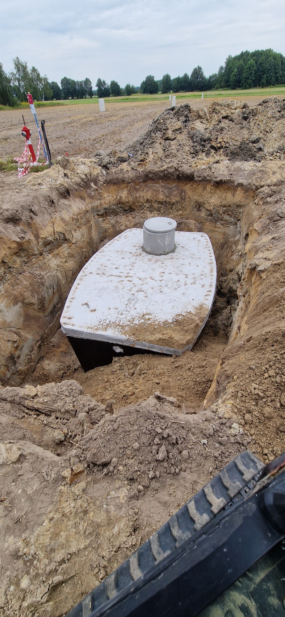 Montaż szamba betonowego w wykopie, widoczne ślady pracy koparki, w tle pole i drzewa.