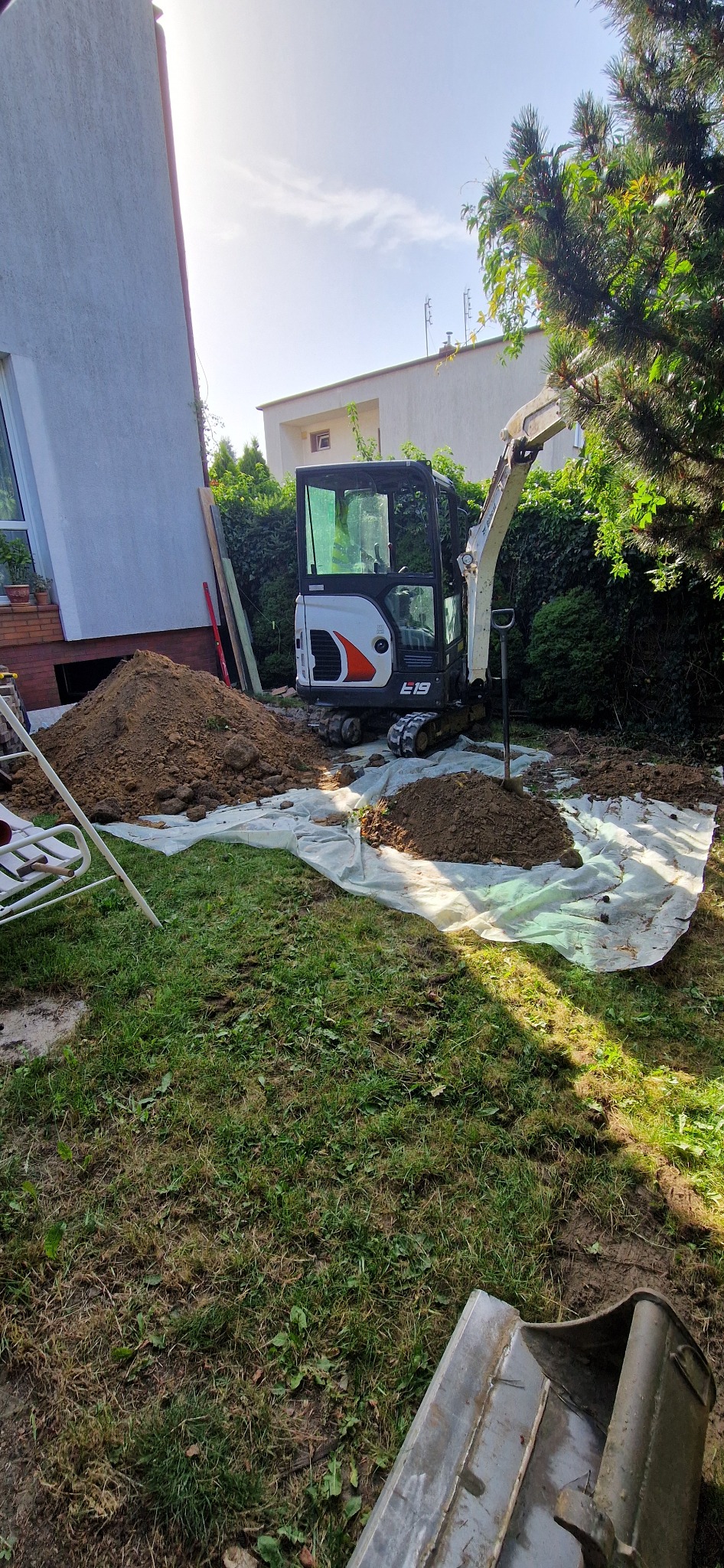 Mała koparka Bobcat E19 na trawniku, z pryzmą ziemi na plandece, widoczny budynek i zieleń w tle.