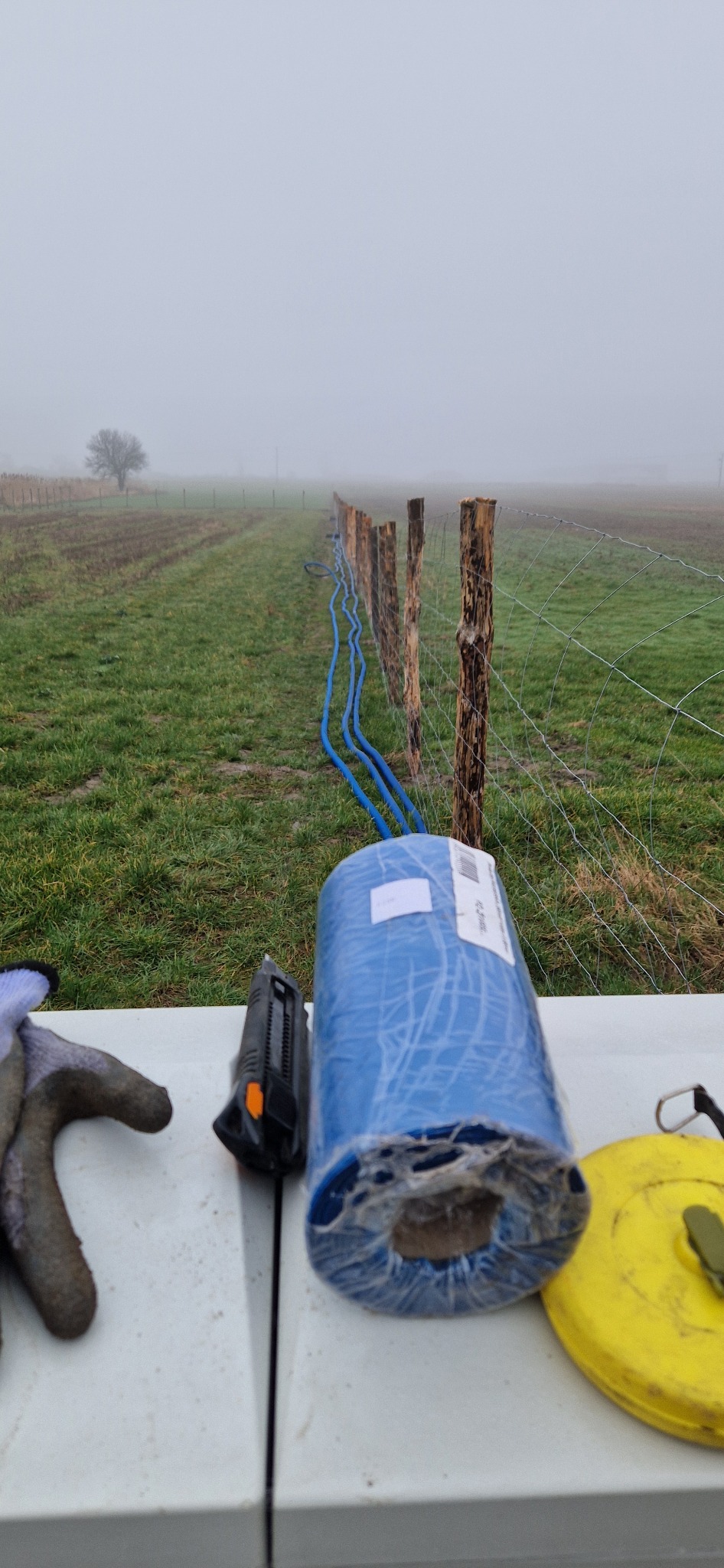 Instalacja niebieskiego przewodu wzdłuż ogrodzenia na zewnątrz w mglisty dzień, widoczny zwój materiału, nóż, rękawica i miarka na stole.