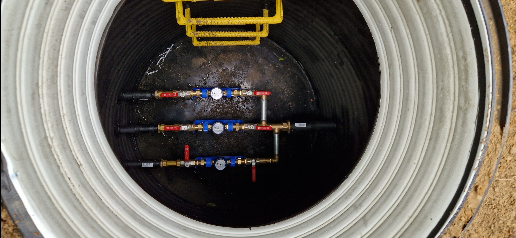 Wnętrze betonowego zbiornika z trzema rzędami instalacji wodociągowych z wodomierzami, zaworami i złączkami, widoczne żółte elementy drabinki.