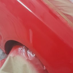 ŁUKASZ - Czerwony błotnik samochodu po lakierowaniu, zabezpieczony folią i papierem maskującym. Widoczne odbicia światła na świeżej powłoce lakierniczej.