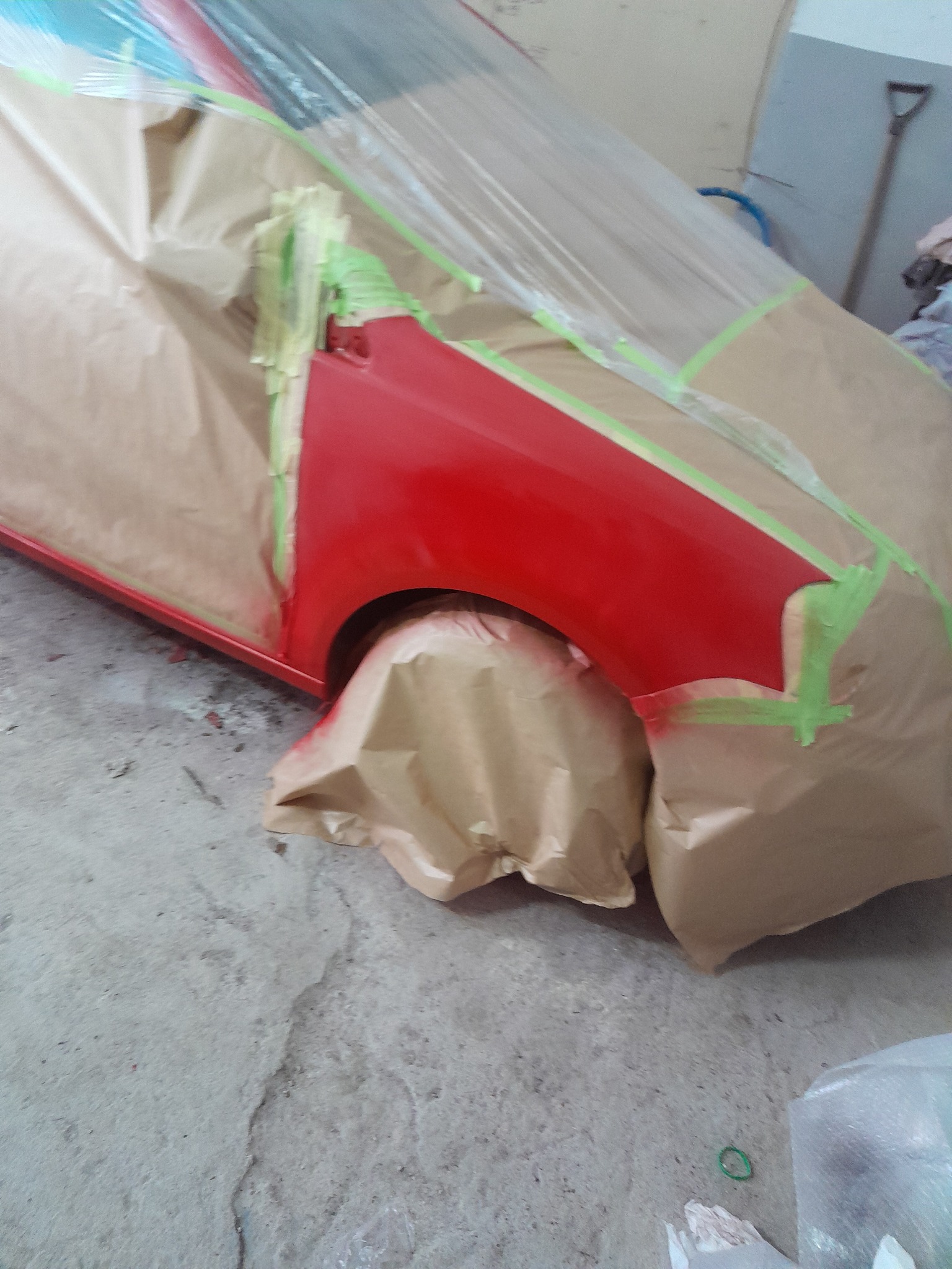 Czerwony samochód oklejony papierem i taśmą malarską, przygotowany do lakierowania. Widoczne fragmenty karoserii i opony. W tle ściana warsztatu i narzędzie.