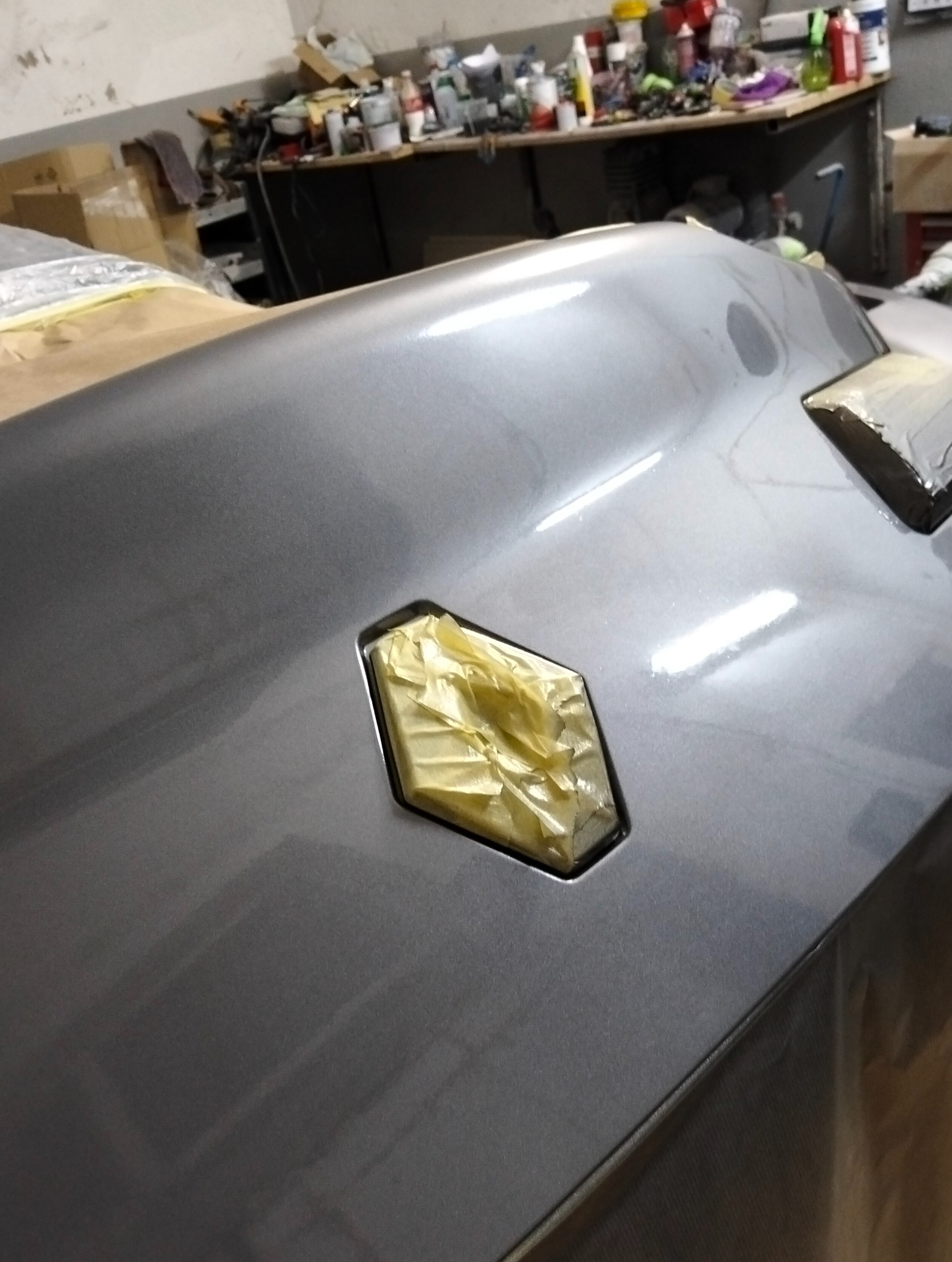 Szary element karoserii auta z zabezpieczonymi taśmą fragmentami, przygotowany do lakierowania. Widoczne refleksy światła na świeżo położonym lakierze.