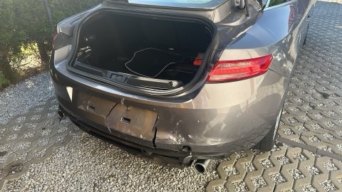 Uszkodzony, szary samochód z otwartym bagażnikiem. Widoczne pęknięcia i wgniecenia na tylnym zderzaku. Auto stoi na kostce brukowej.
