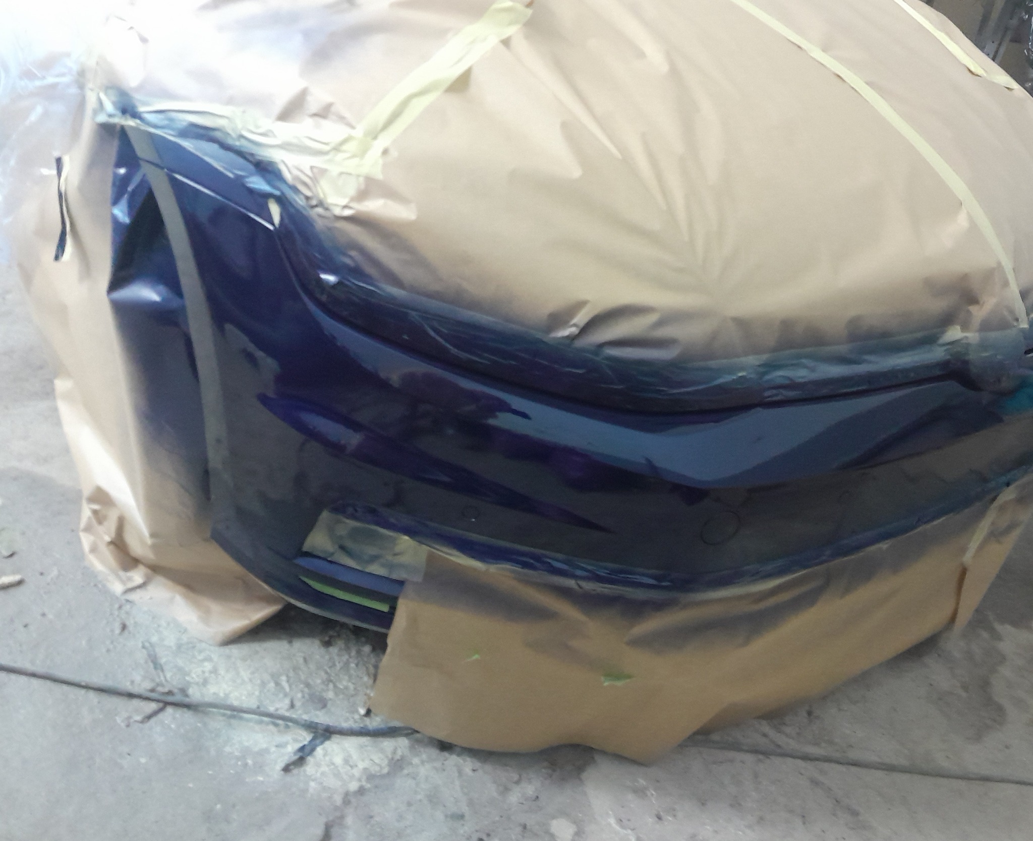 Błyszczący, granatowy zderzak samochodowy, zabezpieczony folią i papierem maskującym przed lakierowaniem, w warsztacie. Detal z bliska, przygotowanie do lakierowania.