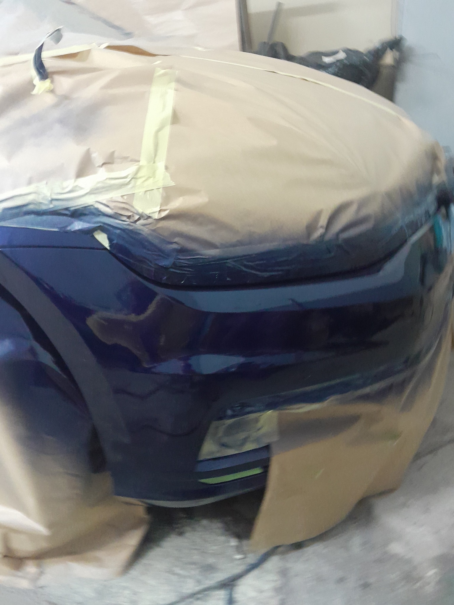 Niebieski błotnik auta, częściowo zakryty papierem i taśmą malarską, przygotowany do lakierowania. Widoczne detale karoserii i oświetlenia.