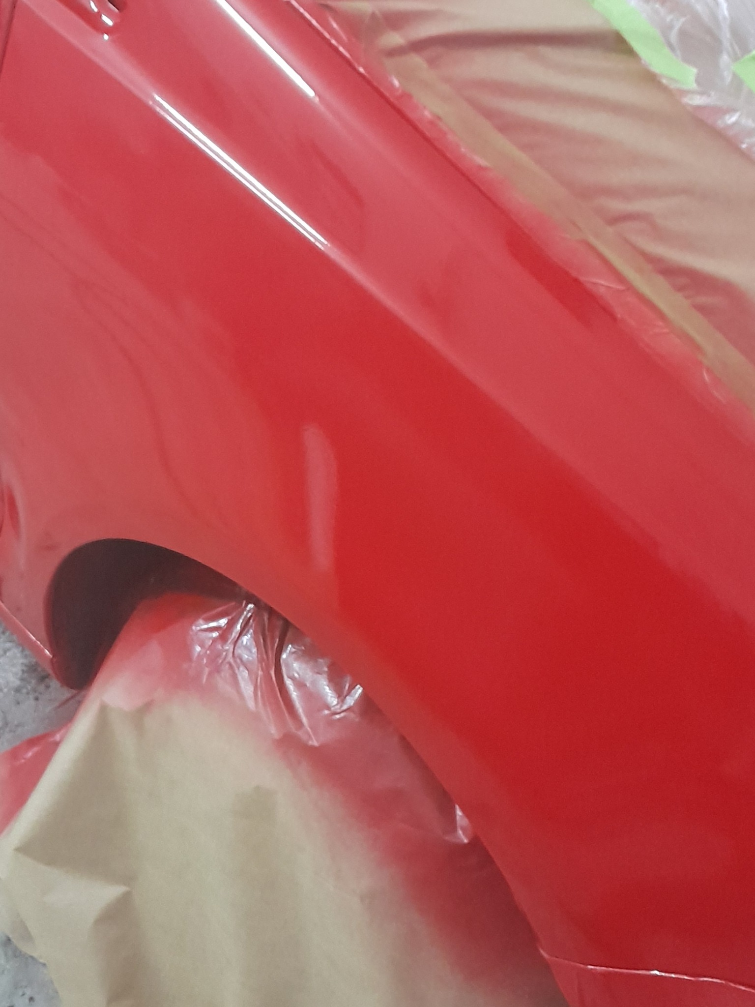 Czerwony błotnik samochodu po lakierowaniu, zabezpieczony folią i papierem maskującym. Widoczne odbicia światła na świeżej powłoce lakierniczej.