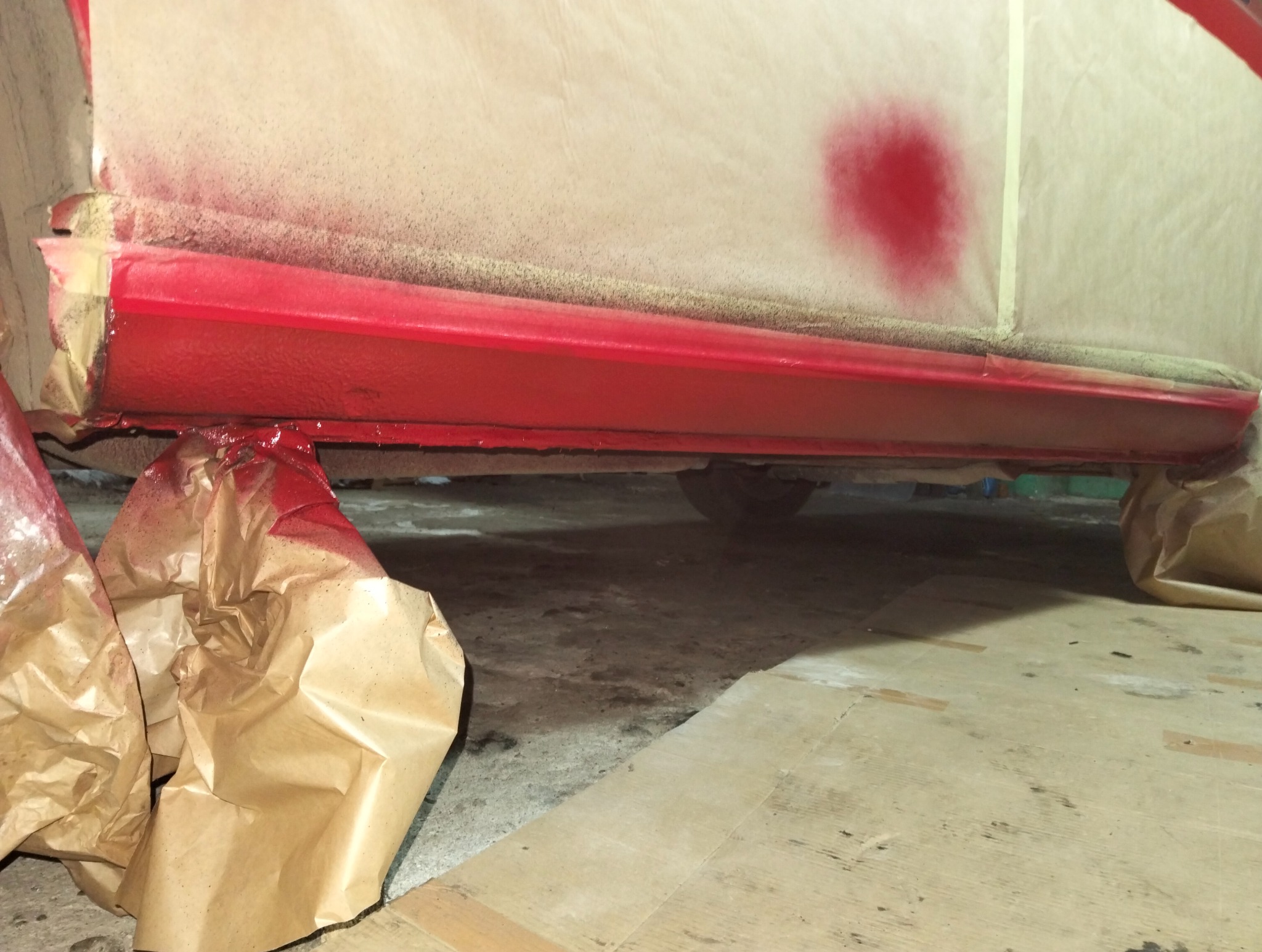 Czerwony próg samochodu w trakcie lakierowania, zabezpieczony papierem i taśmą. Widoczne ślady farby na papierze ochronnym.