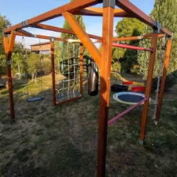 Drewniana konstrukcja placu zabaw w ogrodzie z huśtawką, siatką do wspinaczki, workiem bokserskim i drabinką sznurową. W tle zieleń i budynek.
