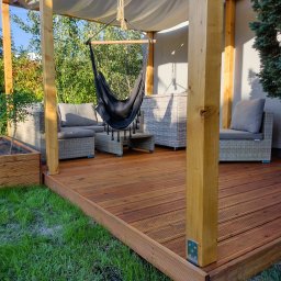 WoodByte - Zadaszony taras z drewna z wiszącym fotelem hamakowym i meblami ogrodowymi z rattanu, widok na zadbany trawnik i zieleń ogrodu.
