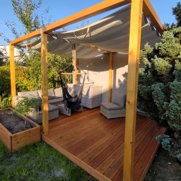 WoodByte - Drewniany taras z jasną, drewnianą pergolą i materiałowym zadaszeniem, wyposażony w rattanowe meble ogrodowe i wiszący fotel hamakowy, otoczony zielenią.