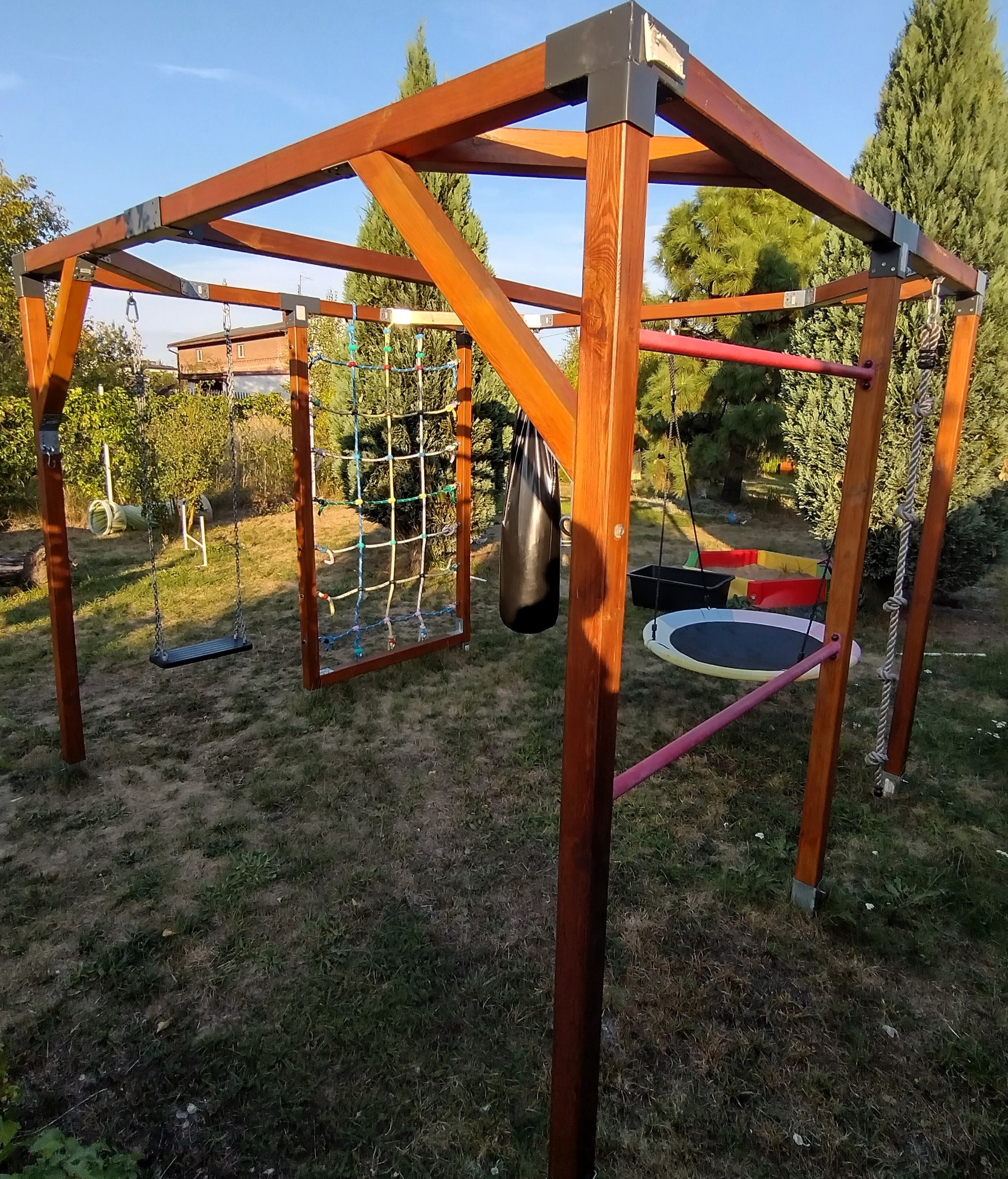 Drewniana konstrukcja placu zabaw w ogrodzie z huśtawką, siatką do wspinaczki, workiem bokserskim i drabinką sznurową. W tle zieleń i budynek.