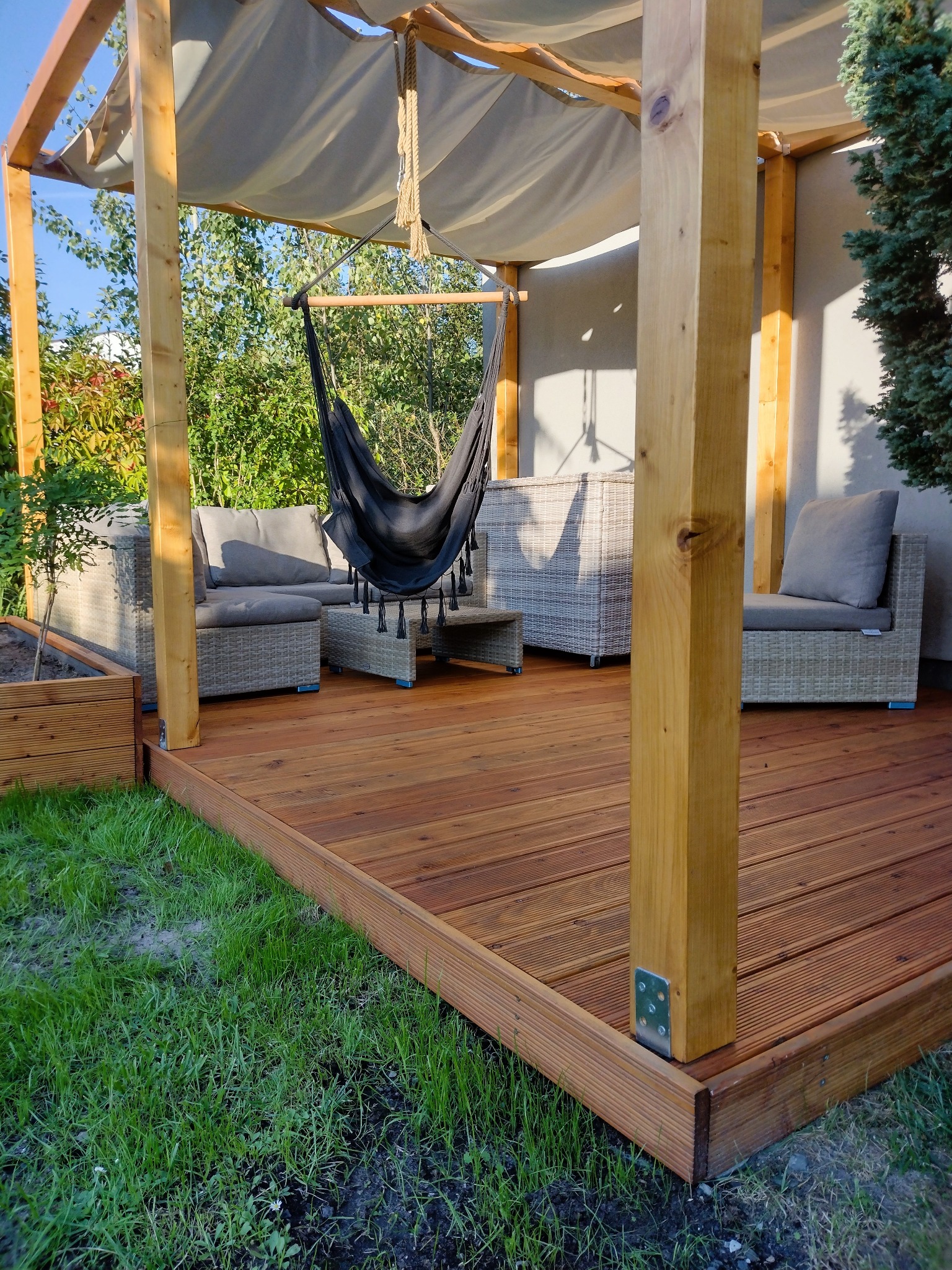 Zadaszony taras z drewna z wiszącym fotelem hamakowym i meblami ogrodowymi z rattanu, widok na zadbany trawnik i zieleń ogrodu.