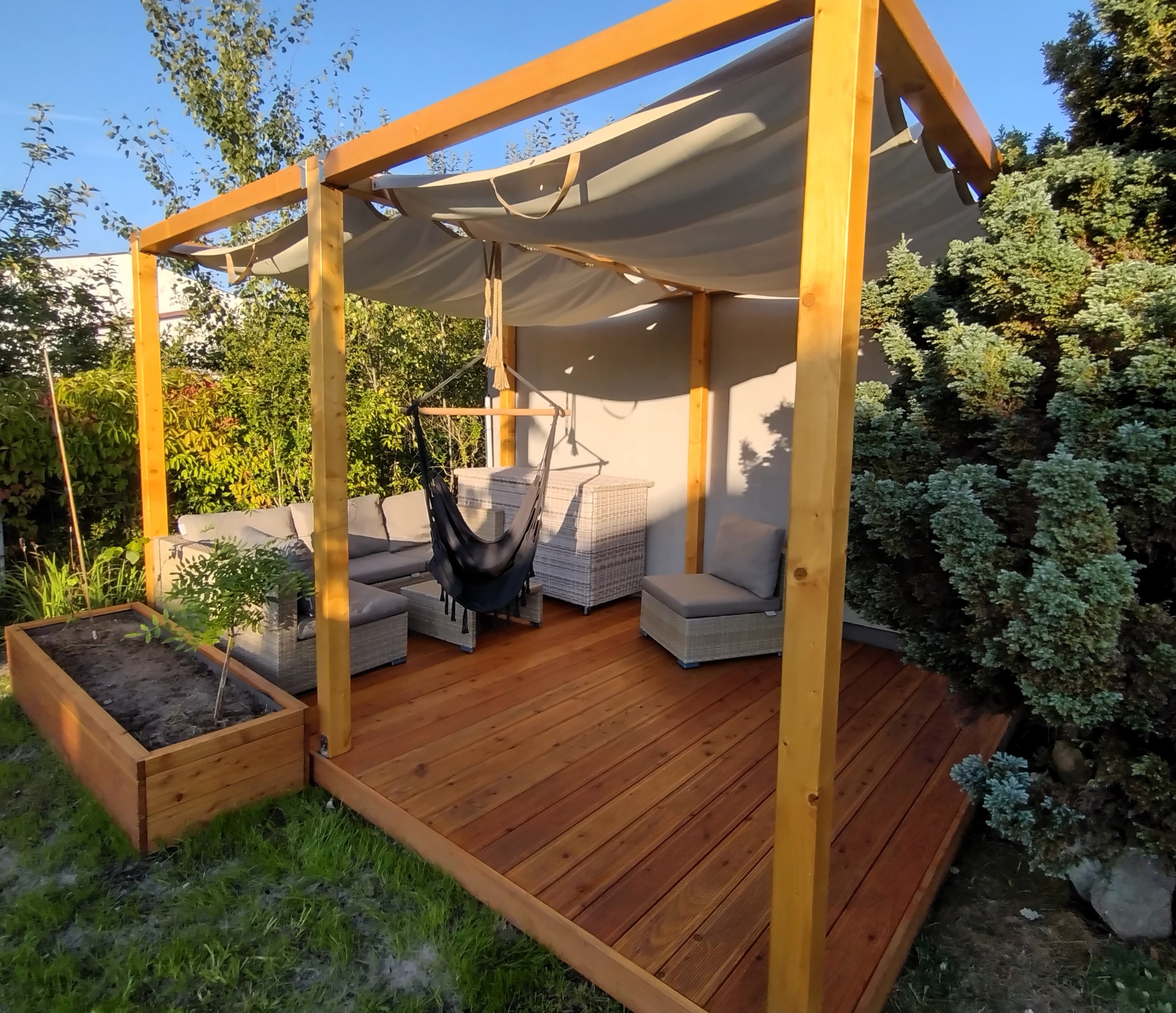 Drewniany taras z jasną, drewnianą pergolą i materiałowym zadaszeniem, wyposażony w rattanowe meble ogrodowe i wiszący fotel hamakowy, otoczony zielenią.