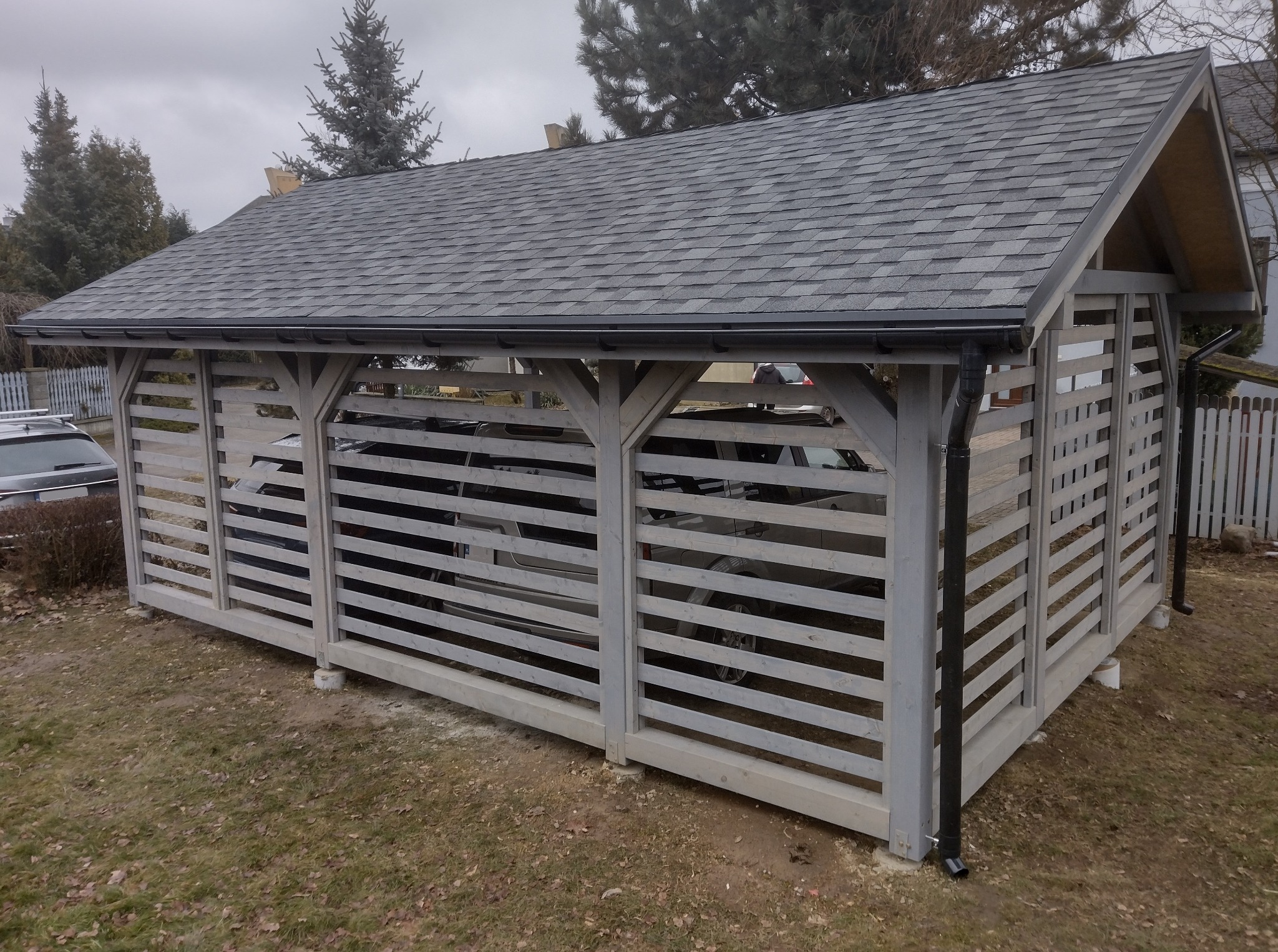 Drewniana wiata garażowa z szarym dachem bitumicznym, osłonięta ażurowymi panelami, z zaparkowanym samochodem wewnątrz, na działce z drzewami w tle.