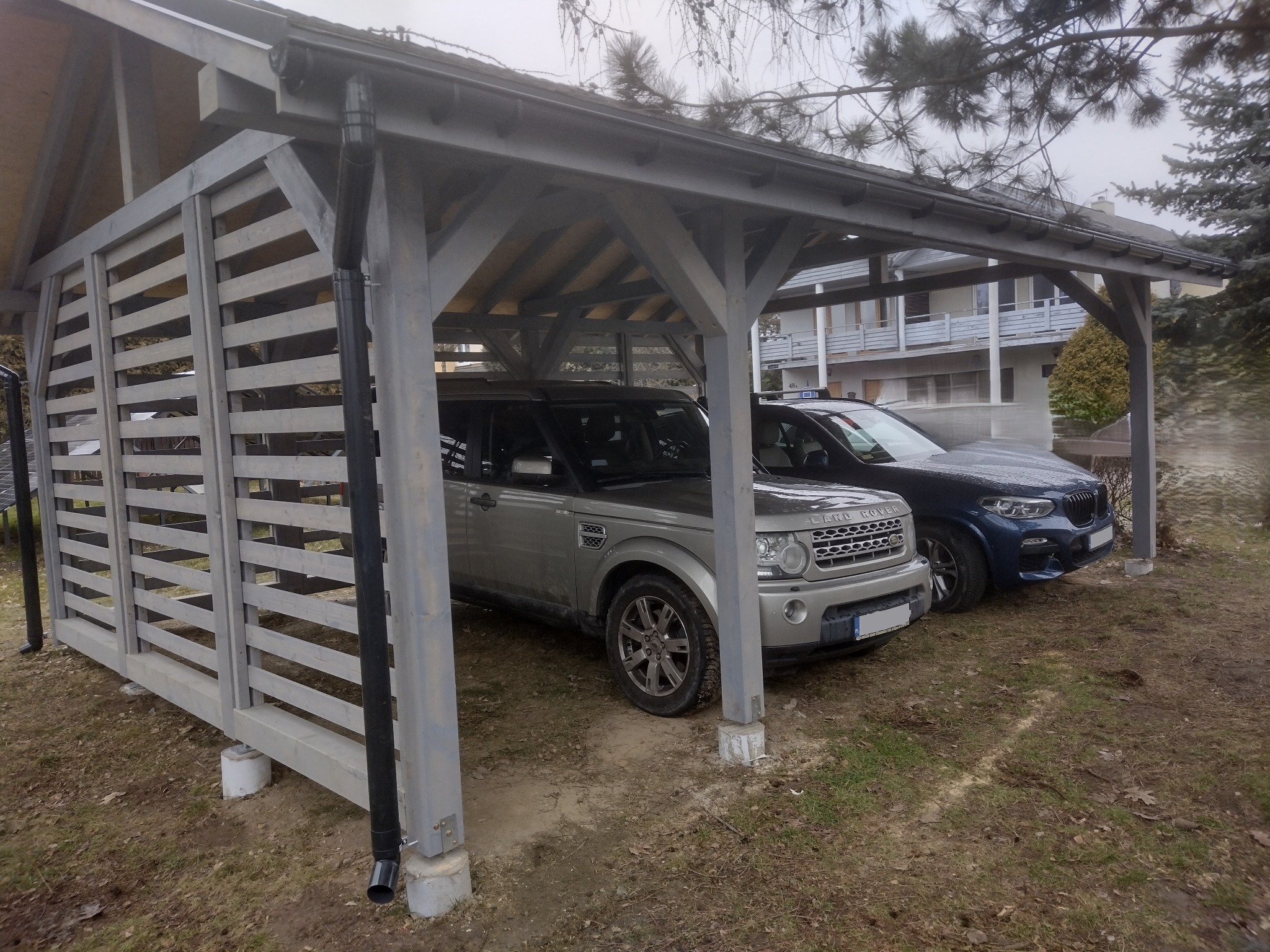 Drewniana wiata garażowa na dwa samochody, z szarymi elementami konstrukcyjnymi i gontem bitumicznym na dachu, osłonięta z jednej strony drewnianymi panelami lamelowymi, z widocznymi dwoma...