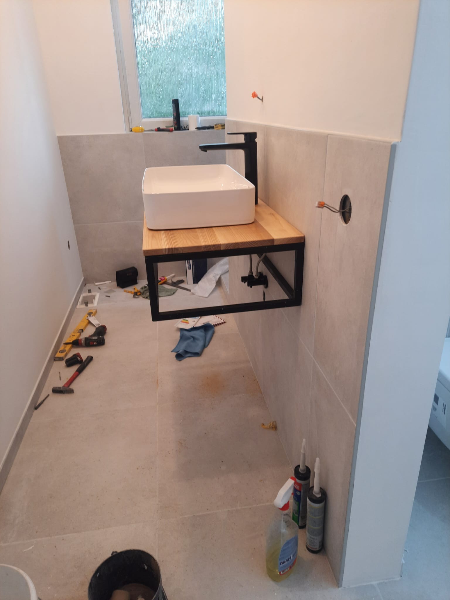 Łazienka w trakcie montażu umywalki nablatowej z czarną armaturą na drewnianym blacie z metalową ramą, widoczne narzędzia, poziomica, wiertarka, wiadro i preparaty do czyszczenia na podłodze...