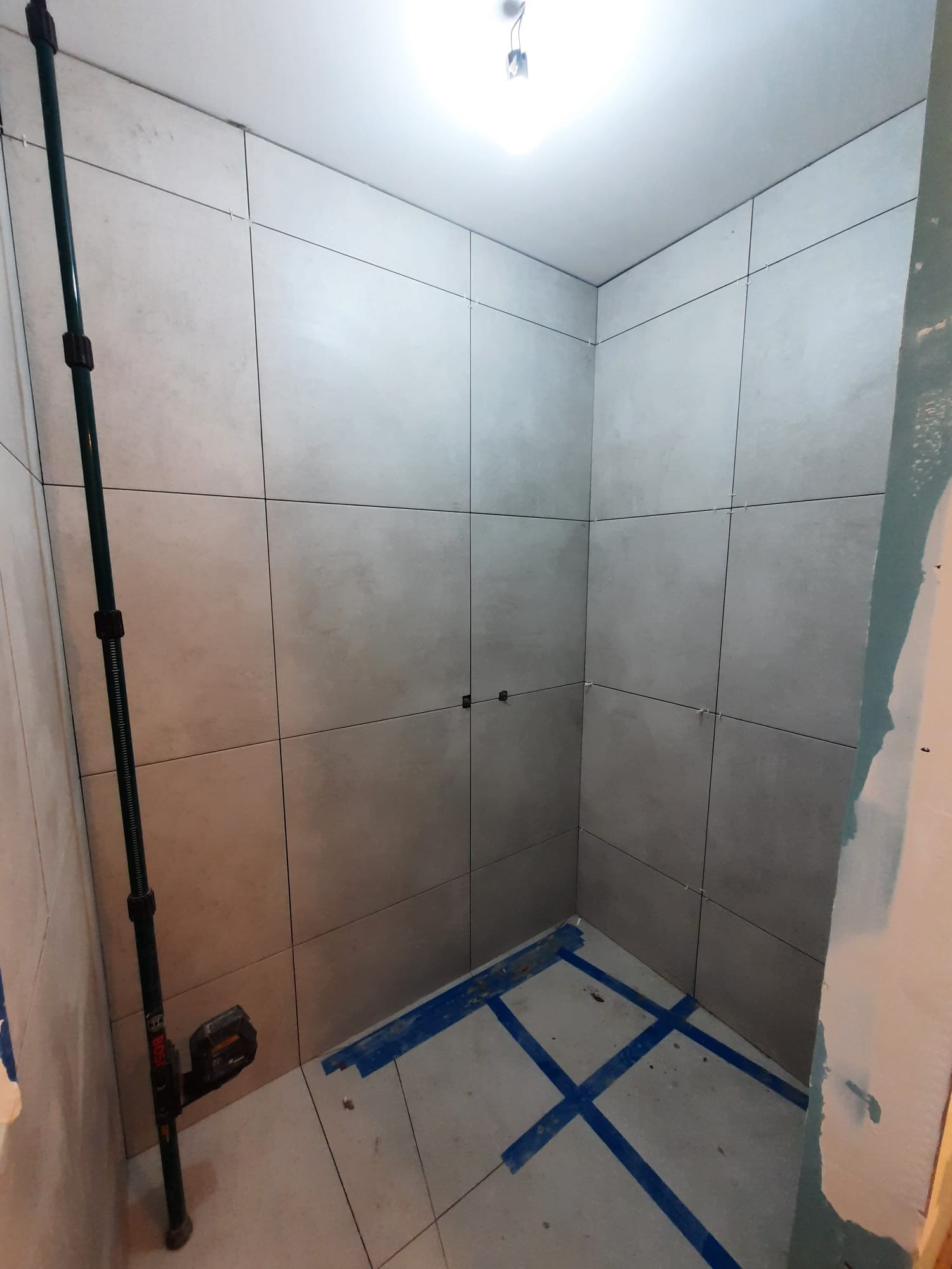 Wnętrze kabiny prysznicowej w trakcie wykańczania, z szarymi płytkami na ścianach i podłodze, poziomica laserowa Bosch ustawiona w rogu, niebieska taśma wyznaczająca linie na podłodze, widoczne...