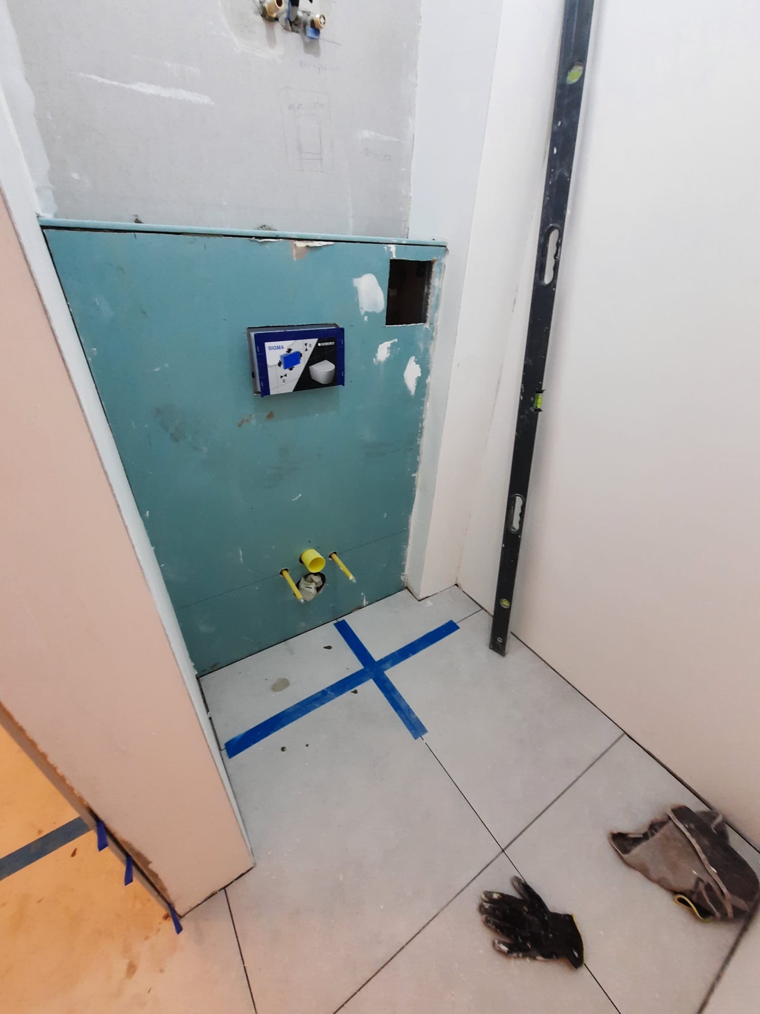 Przygotowania do montażu podtynkowego zestawu WC w łazience w Warszawie. Widoczne niebieskie taśmy wyznaczające układ płytek, poziomica oparta o ścianę, oraz wystające rury instalacji wodnej.