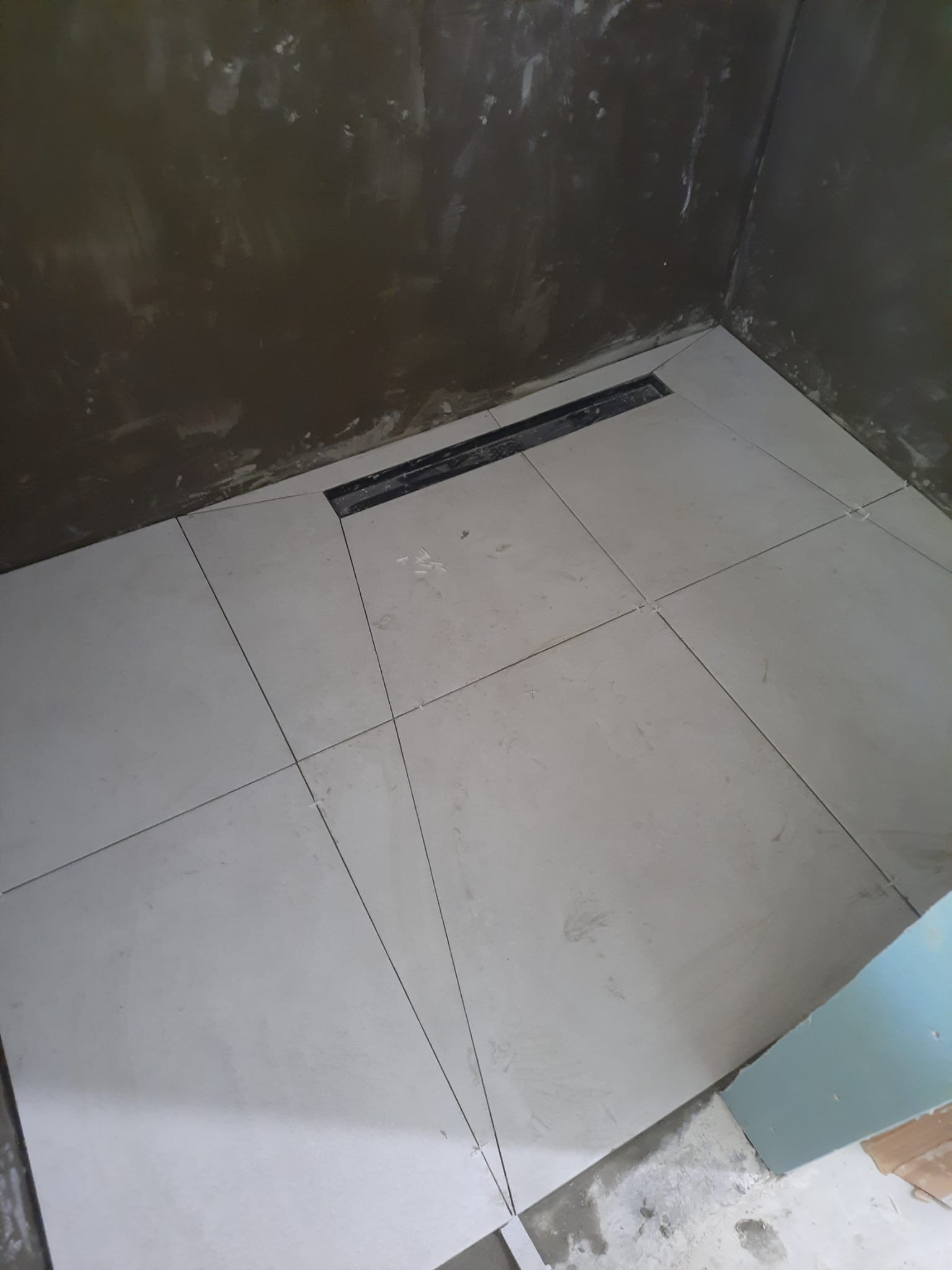 Ułożone jasne, kwadratowe płytki podłogowe w rogu pomieszczenia, z widocznym odpływem liniowym i krzyżykami dystansowymi, ściany w stanie surowym.