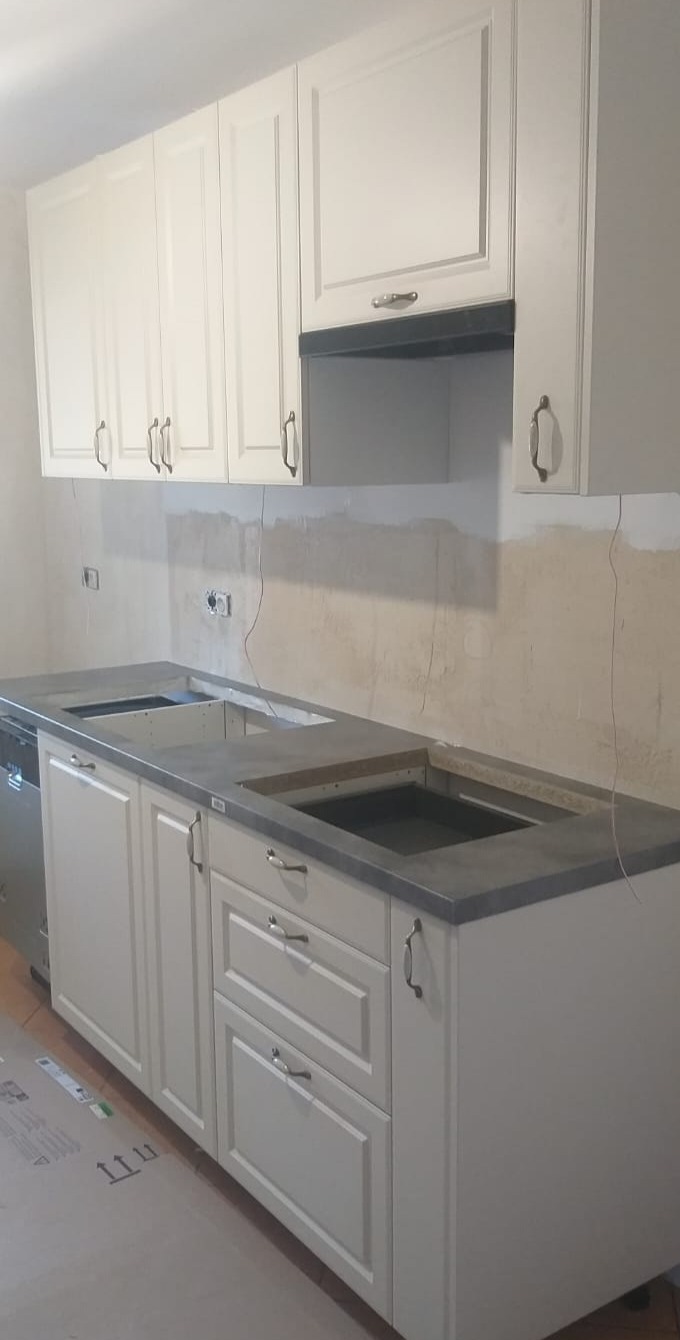 Montaż białych szafek kuchennych z szarym blatem, widoczne otwory na zlew i płytę grzewczą, ściana przygotowana pod płytki.