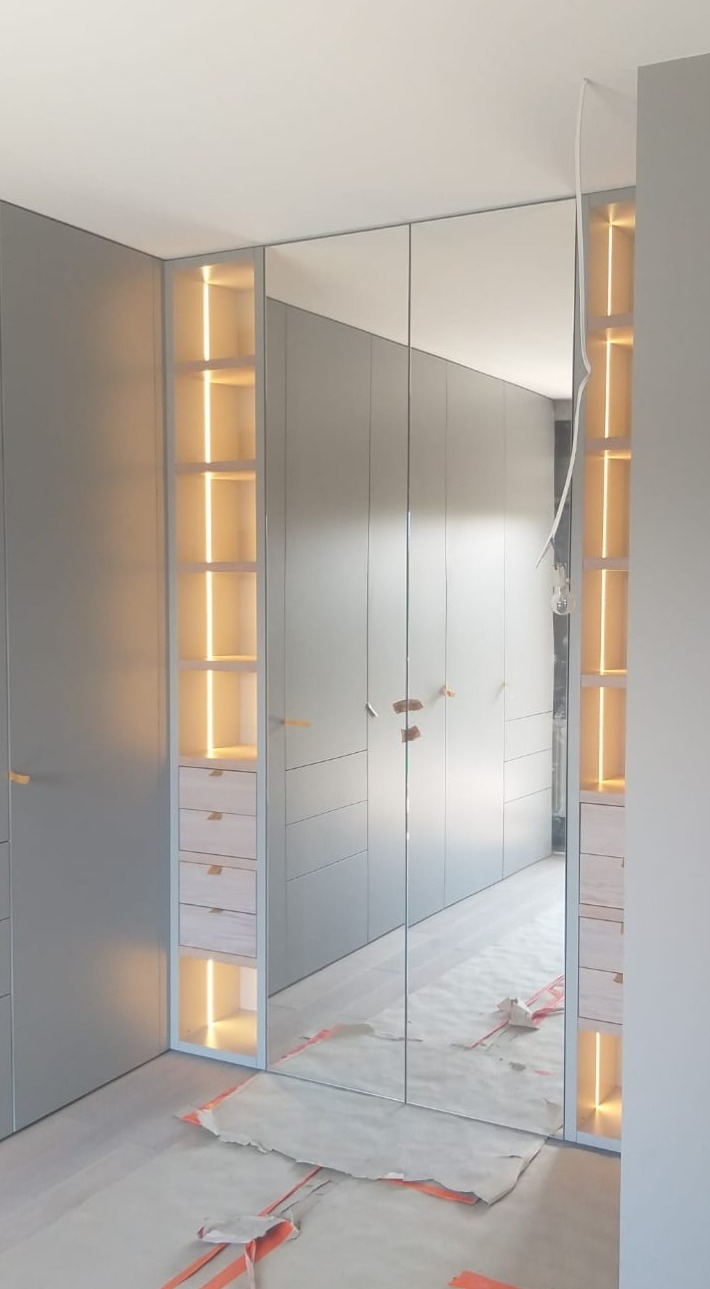 Szara zabudowa szafy z lustrzanymi drzwiami i podświetlanymi półkami, zabezpieczona folią podłoga podczas montażu.