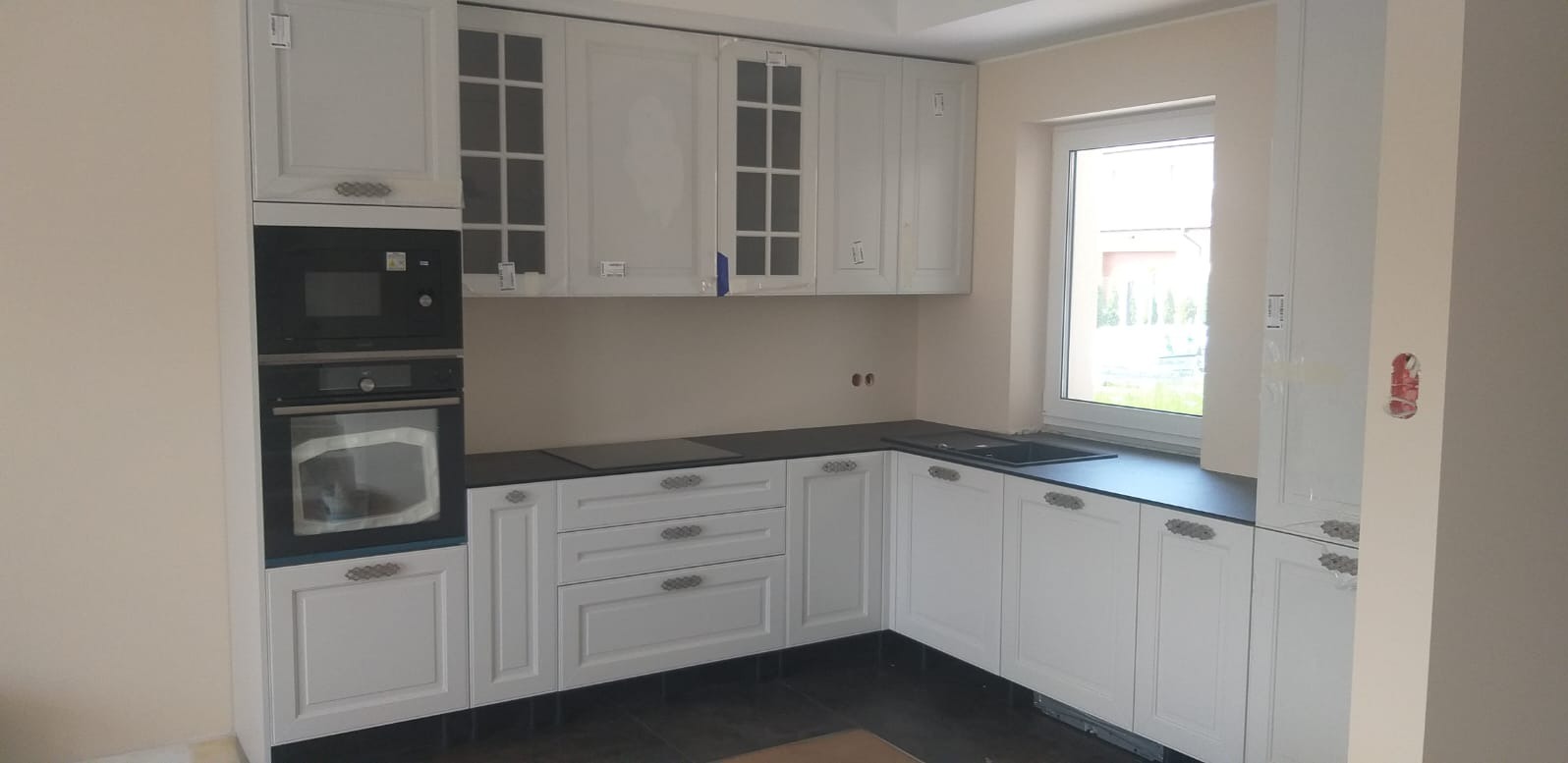 Jasna kuchnia z białymi frezowanymi szafkami i czarnym blatem, wbudowany piekarnik i płyta indukcyjna, widoczne zabezpieczenia folią na szafkach wiszących, ciemna podłoga.