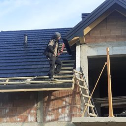 Kamil Pociask - Pracownik w odzieży roboczej montuje obróbkę blacharską na krawędzi nowo położonego, ciemnego dachu z dachówki ceramicznej na surowym budynku z cegły.