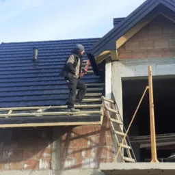 Pracownik w odzieży roboczej montuje obróbkę blacharską na krawędzi nowo położonego, ciemnego dachu z dachówki ceramicznej na surowym budynku z cegły.