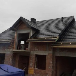 Kamil Pociask - Dom w trakcie budowy z czarnym dachem z dachówki ceramicznej, widoczne murowane ściany z czerwonej cegły, drewniane elementy konstrukcyjne dachu i niebieska plandeka na pierwszym planie.