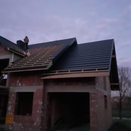 Kamil Pociask - Budowa domu jednorodzinnego w trakcie, widoczne murowane ściany i częściowo pokryty dach z czarnej dachówki, na dachu pracuje dekarz przy kominie, widok o zmierzchu.