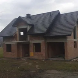 Niedokończony dom jednorodzinny z ciemną dachówką, widoczna murowana konstrukcja ścian i nieotynkowane ściany szczytowe, na pierwszym planie betoniarka.