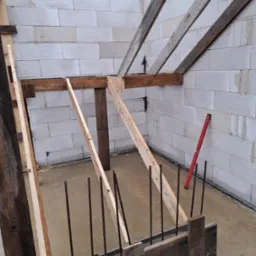 Wykop pod budynek z drewnianym szalunkiem, wystającymi prętami zbrojeniowymi i ścianami z białych bloczków, poziomica oparta o ścianę.