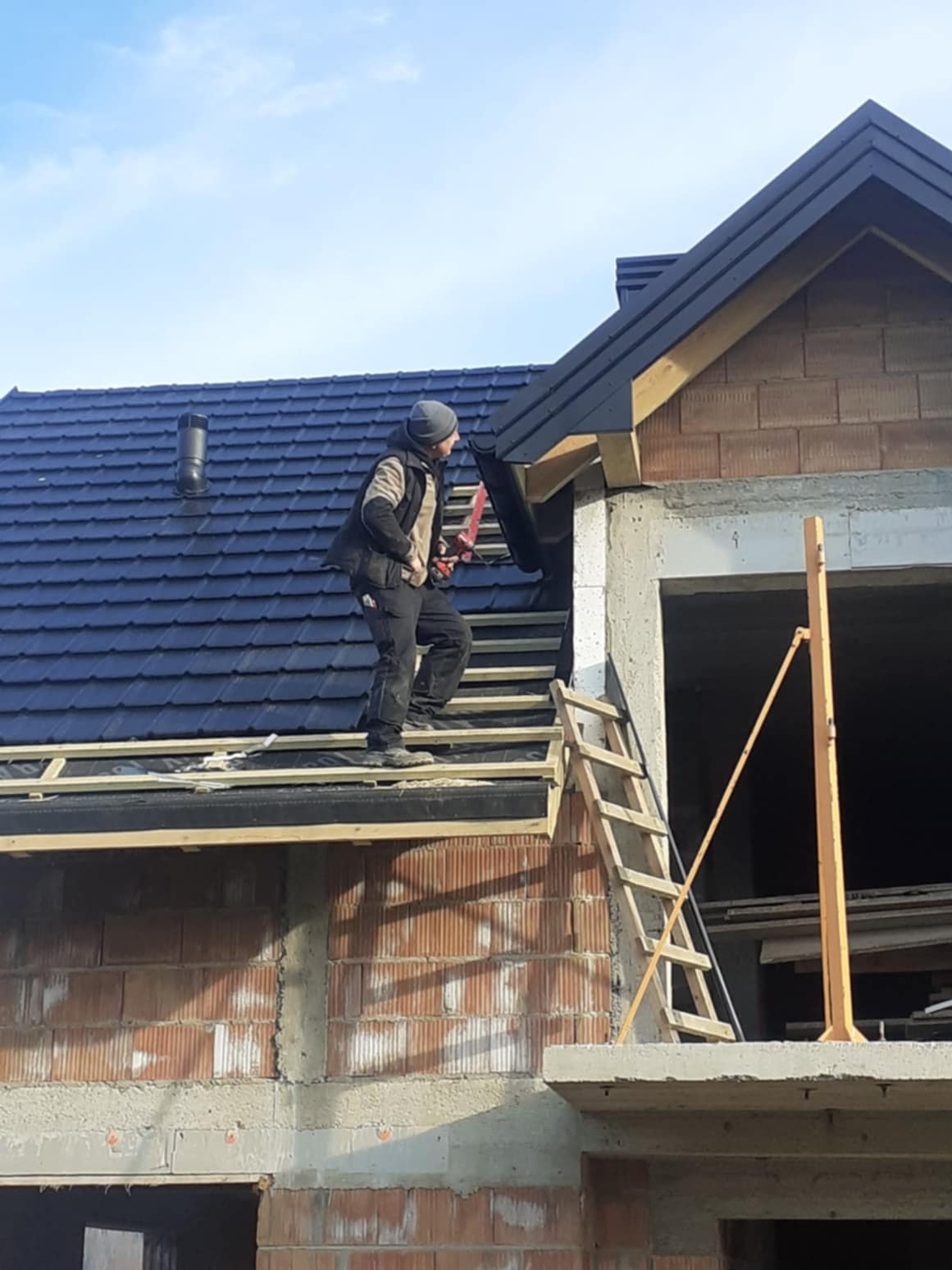 Pracownik w odzieży roboczej montuje obróbkę blacharską na krawędzi nowo położonego, ciemnego dachu z dachówki ceramicznej na surowym budynku z cegły.