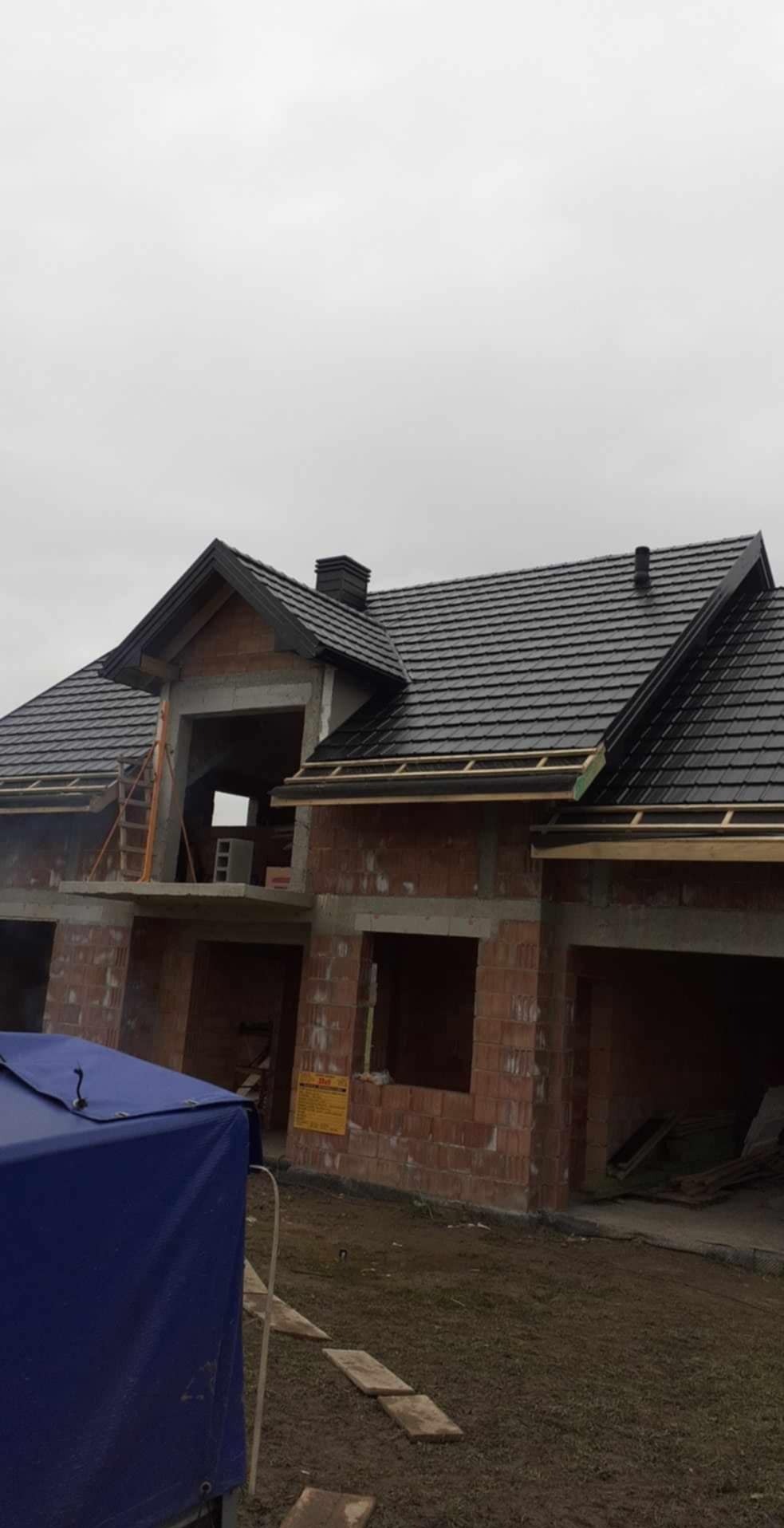 Dom w trakcie budowy z czarnym dachem z dachówki ceramicznej, widoczne murowane ściany z czerwonej cegły, drewniane elementy konstrukcyjne dachu i niebieska plandeka na pierwszym planie.