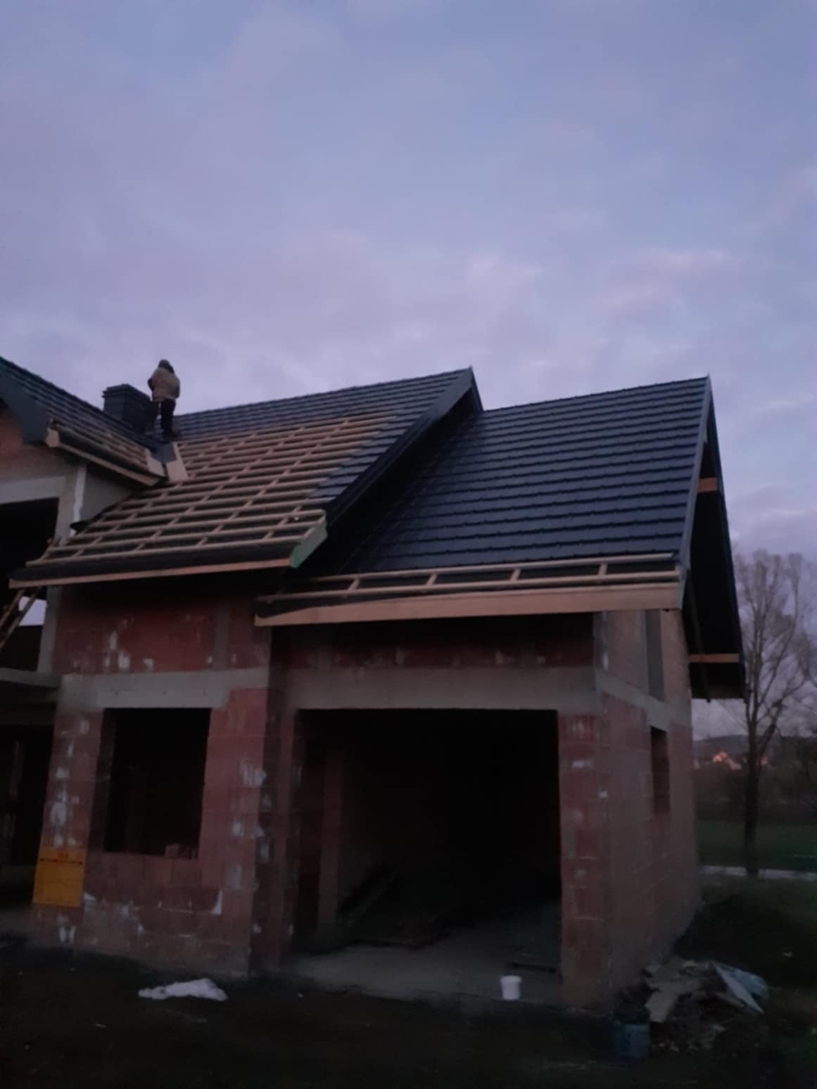 Budowa domu jednorodzinnego w trakcie, widoczne murowane ściany i częściowo pokryty dach z czarnej dachówki, na dachu pracuje dekarz przy kominie, widok o zmierzchu.
